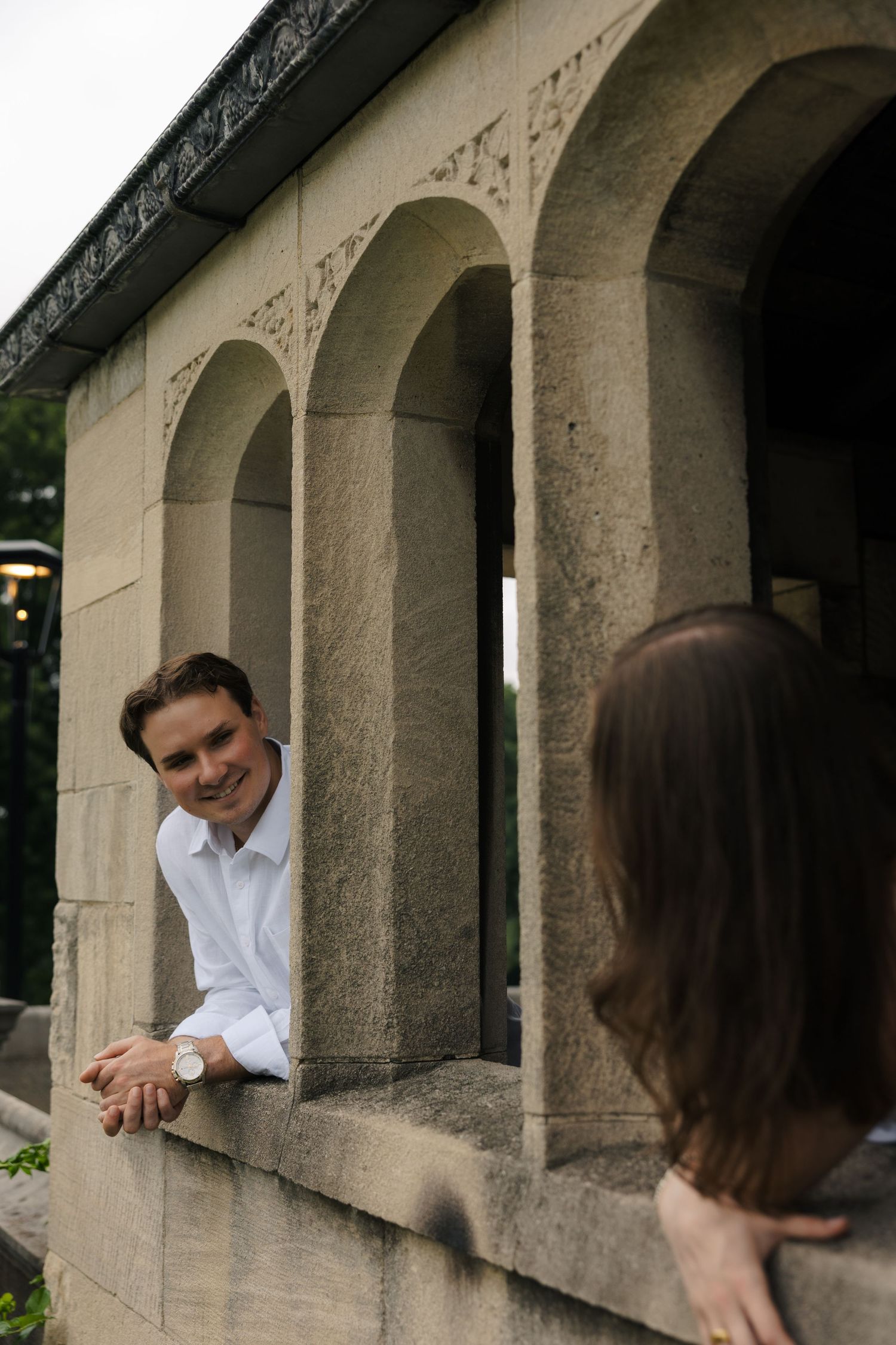 Smiling man in white shirt leaning on stone archway wall looking at woman during engagement photo session.