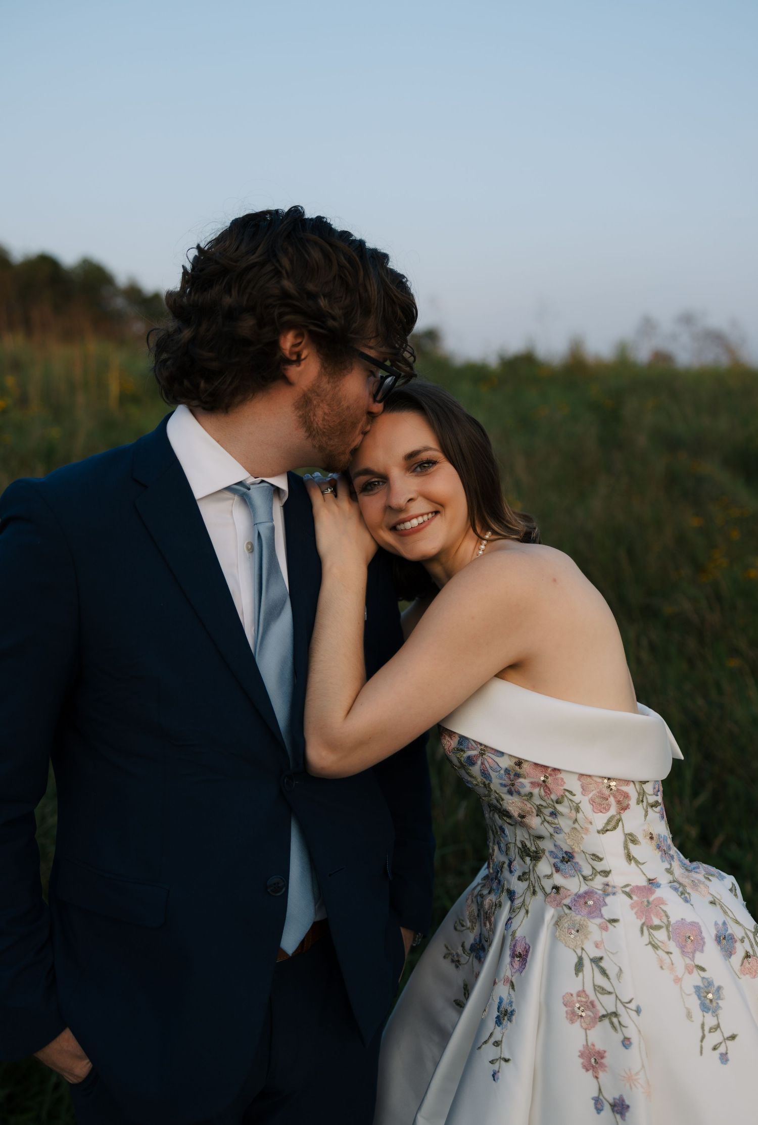 Groom in navy suit kisses bride's cheek as she smiles in floral off-shoulder wedding gown in a field.