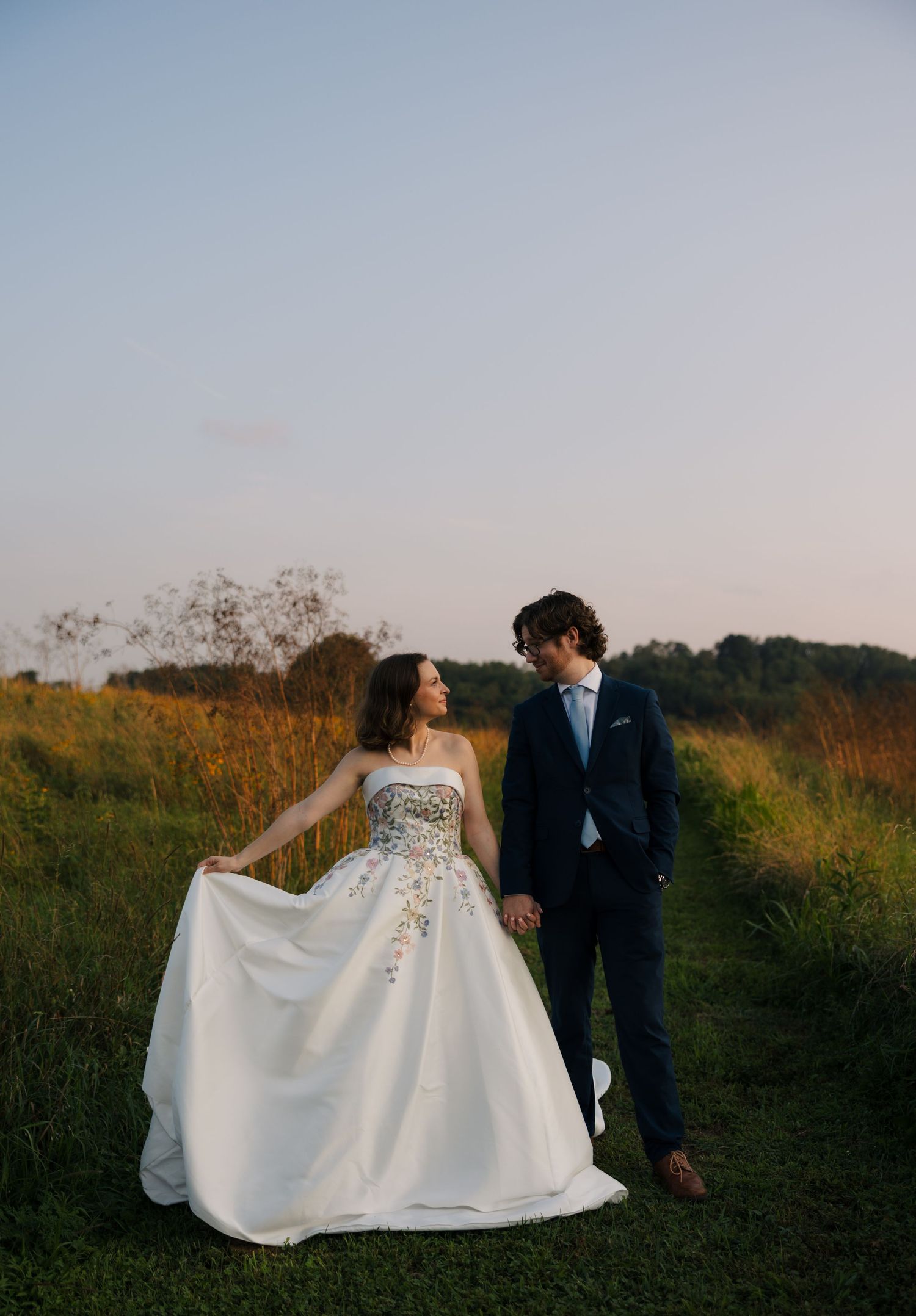 Bride in floral wedding gown holds hands with groom in navy suit walking through green meadow at golden hour.