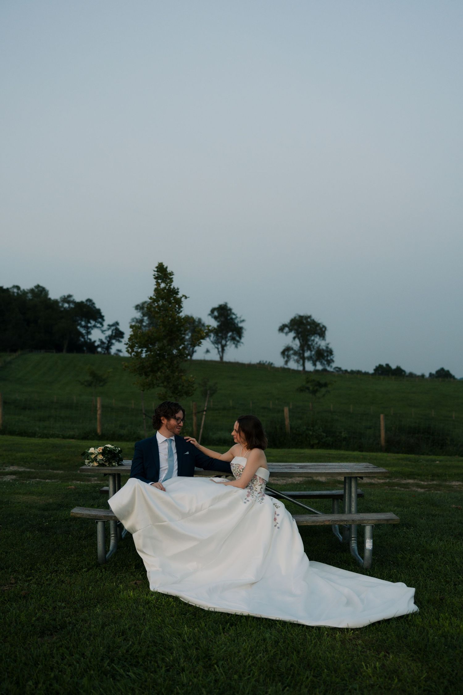 Bride and groom sit together on a picnic bench at dusk, her embroidered gown spread across the green lawn.