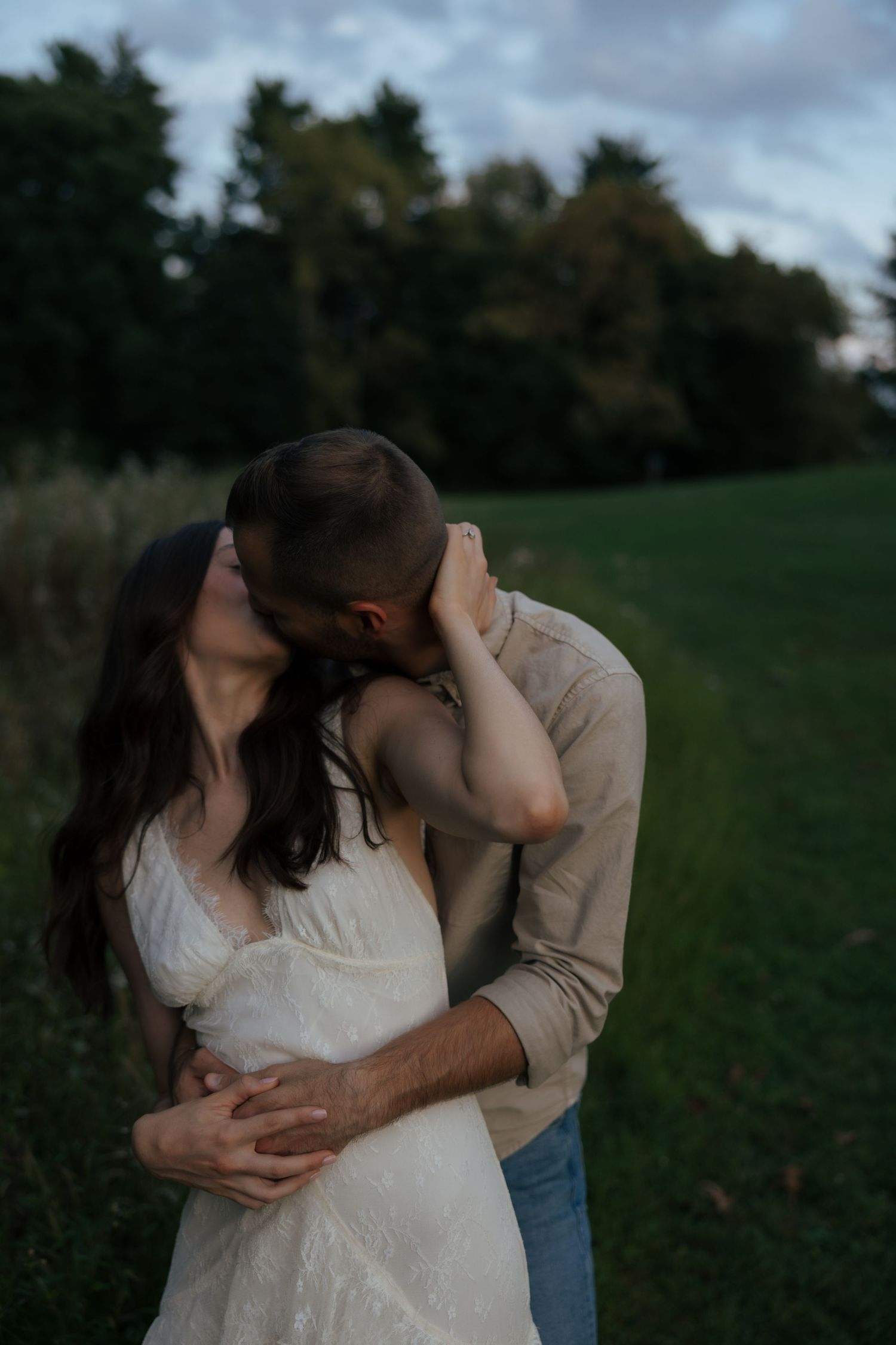 A couple shares a romantic kiss at dusk in a green field, woman in white dress.