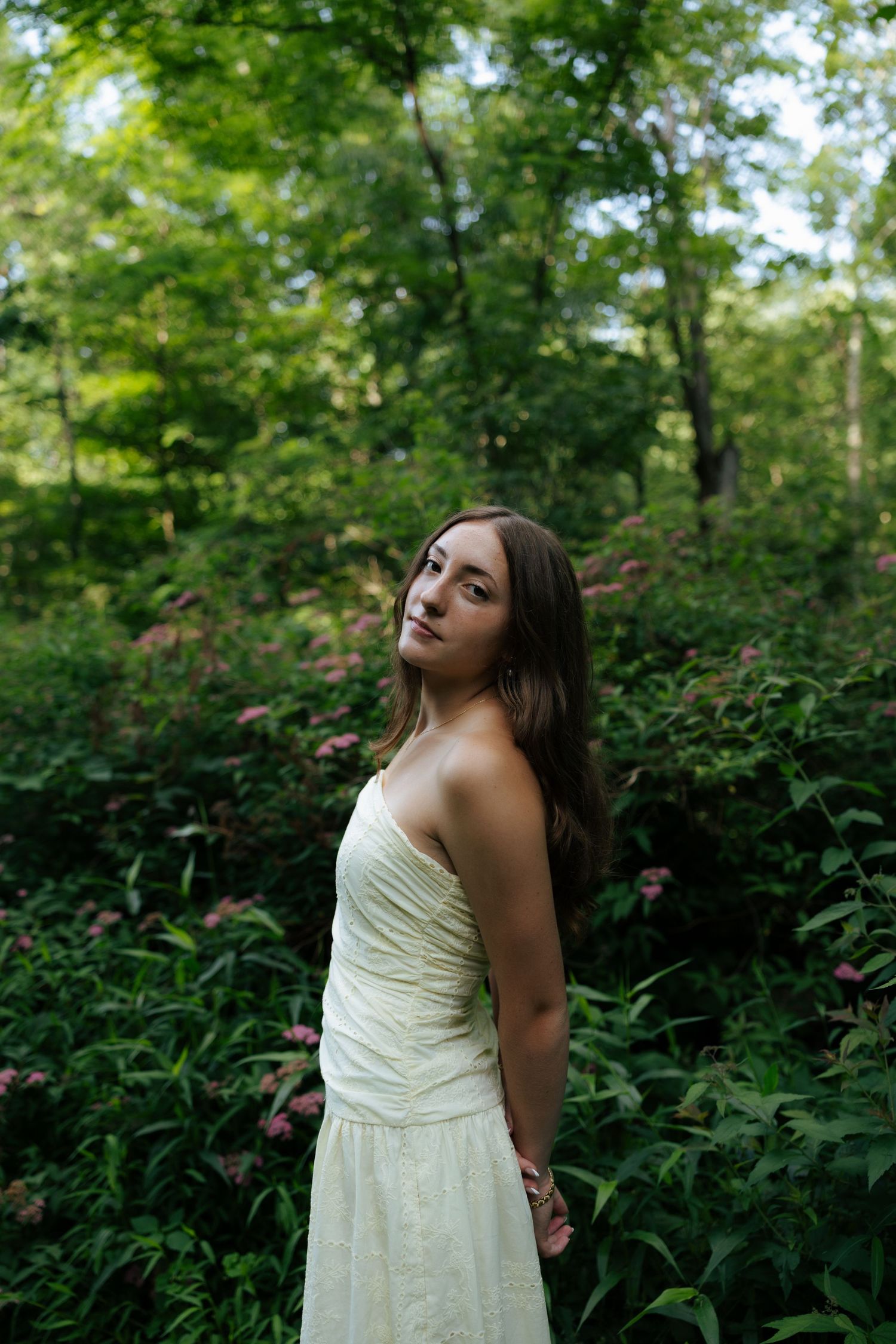 Young brunette woman in cream strapless dress standing among lush green foliage and pink flowers.