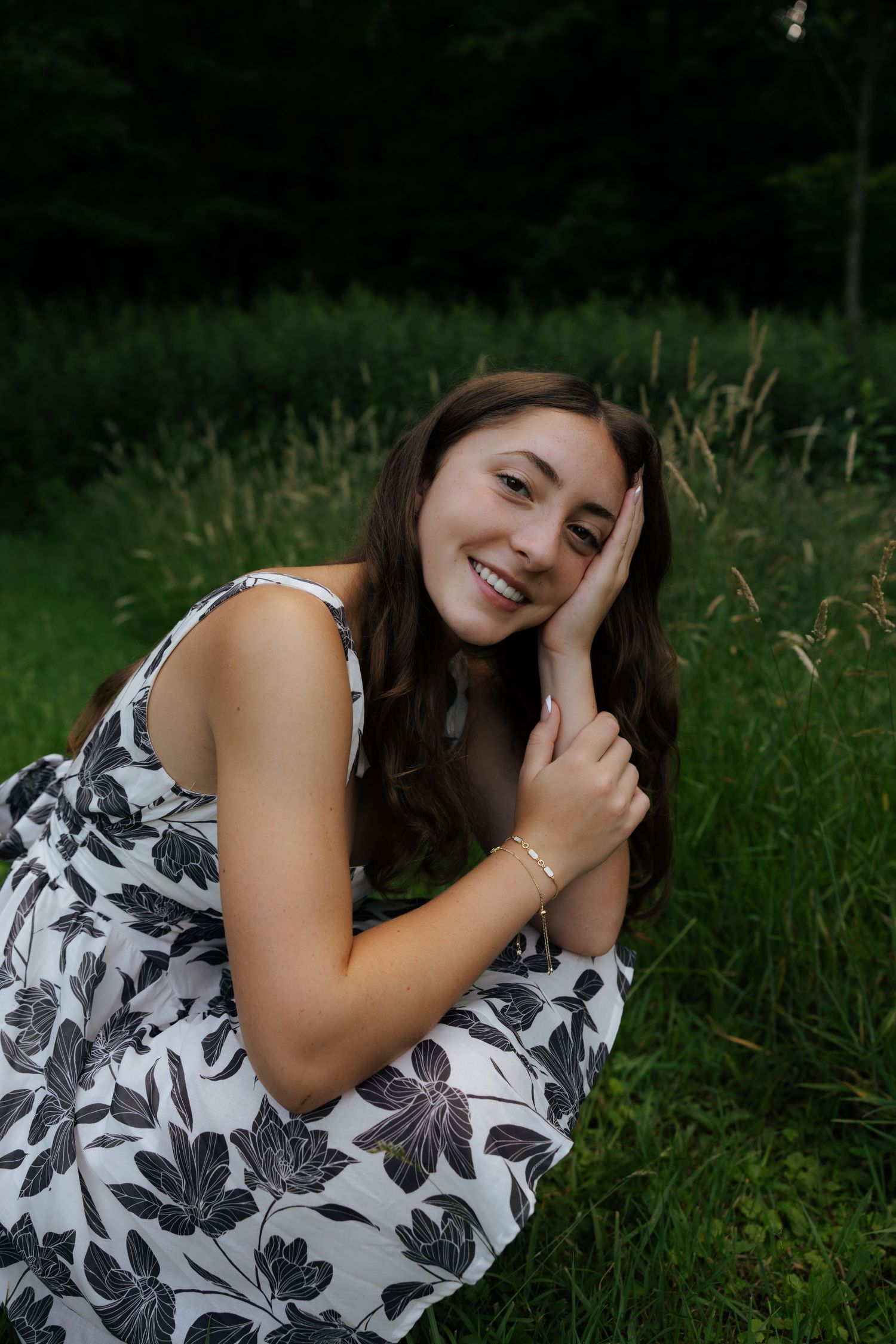 Laughing young woman in floral sundress sitting in grass, hand to face, joyful outdoor portrait.