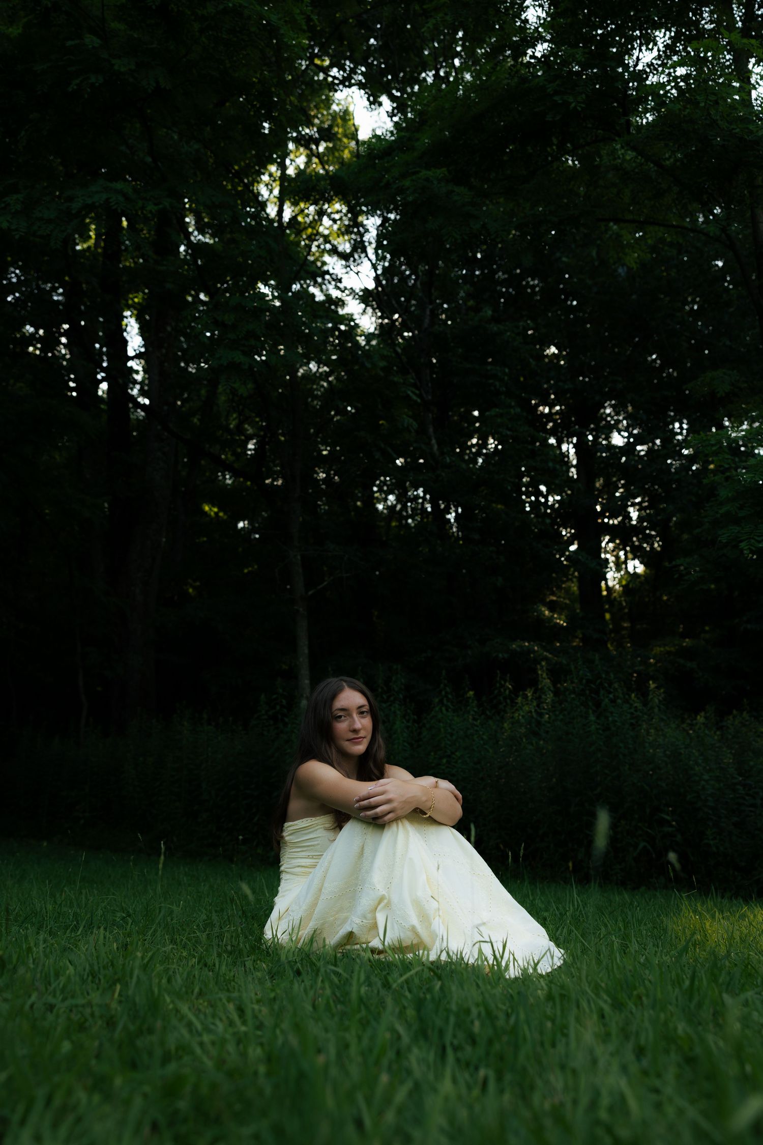 Woman in elegant white dress sitting on lush green grass surrounded by dark forest trees.
