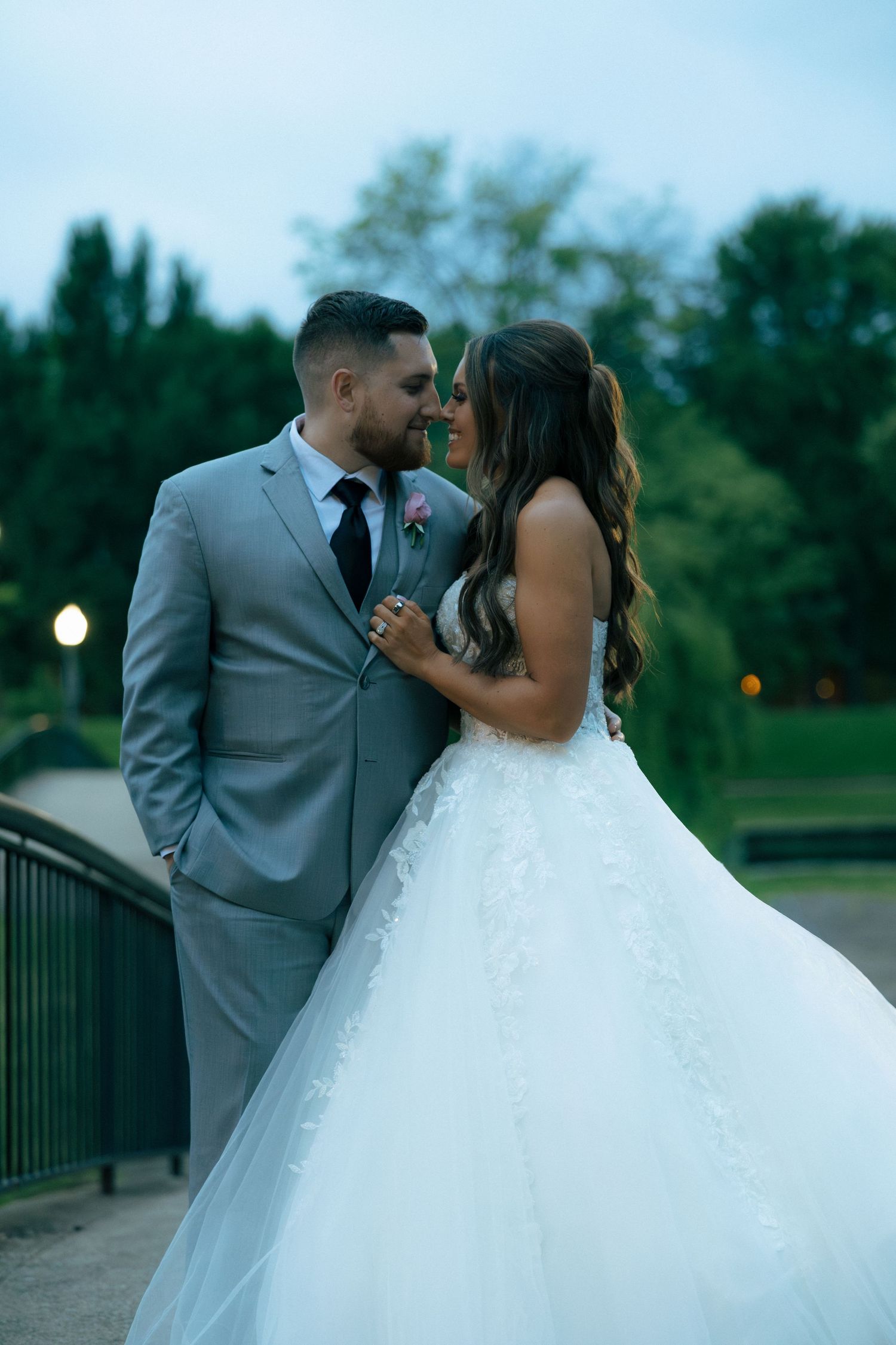 Bride and groom share an intimate moment on a bridge at dusk, bride in a lace ballgown and groom in a gray suit.
