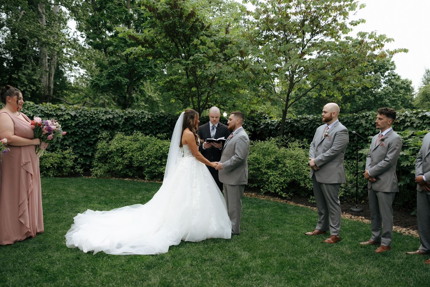 Bride and groom exchange vows at outdoor garden wedding ceremony with bridal party and officiant.