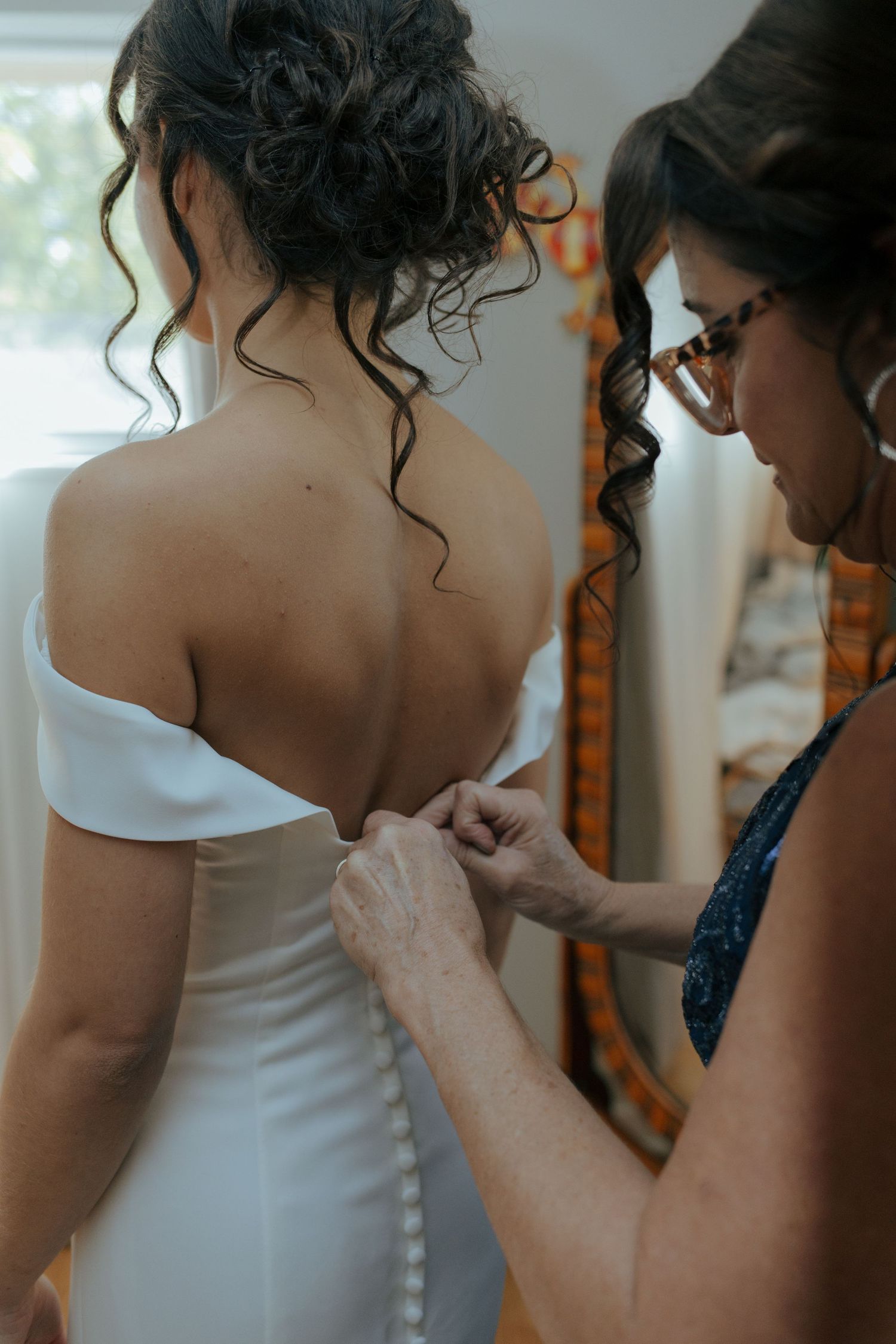 Wedding helper buttoning the back of bride's off-shoulder white satin gown with button detail.