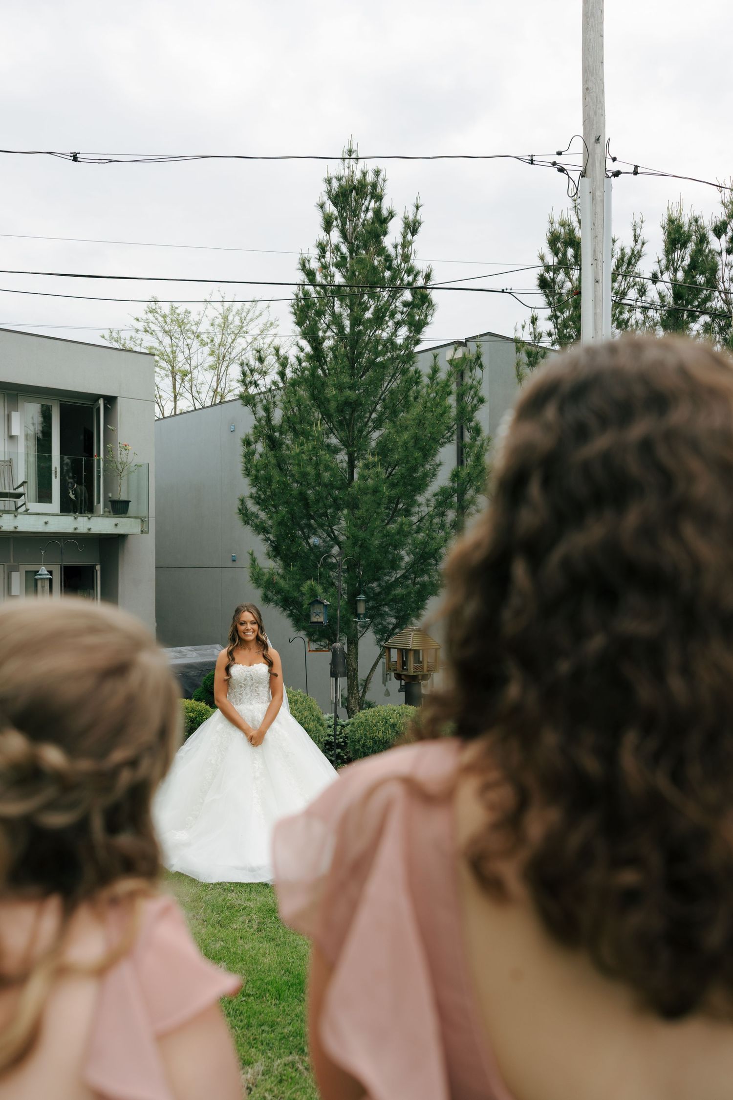 Bride in white ballgown stands on green lawn as bridesmaids in pink dresses look on during outdoor wedding.