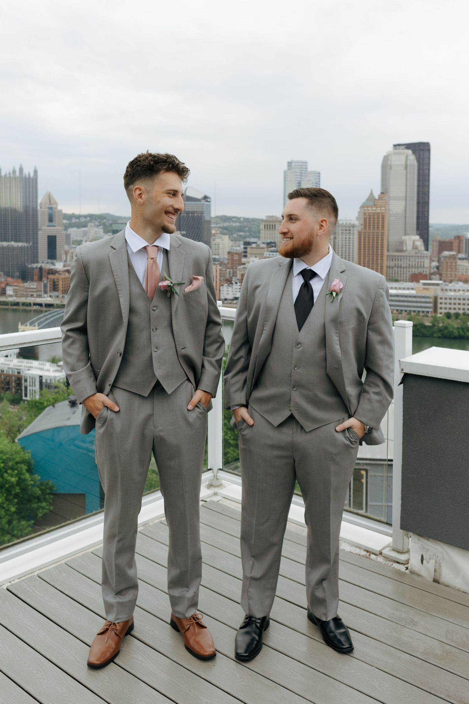 Two grooms in matching gray suits stand on a rooftop with Pittsburgh skyline backdrop on their wedding day.