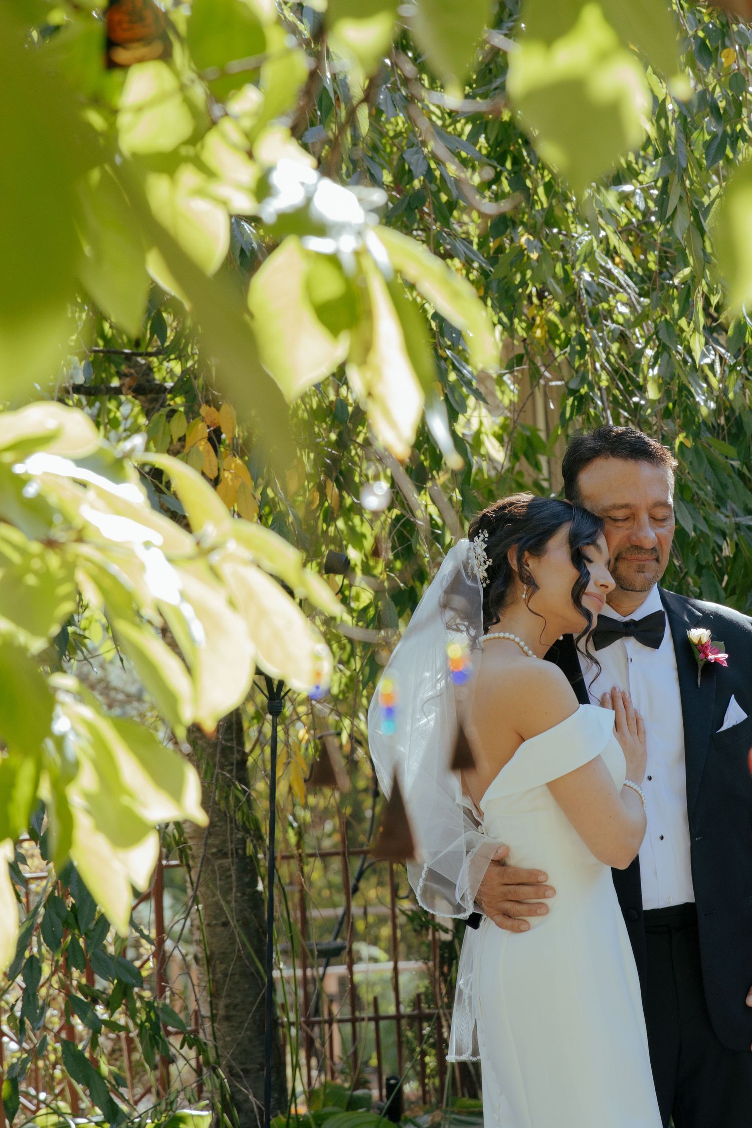Bride and groom share romantic embrace surrounded by lush green foliage during outdoor wedding celebration.