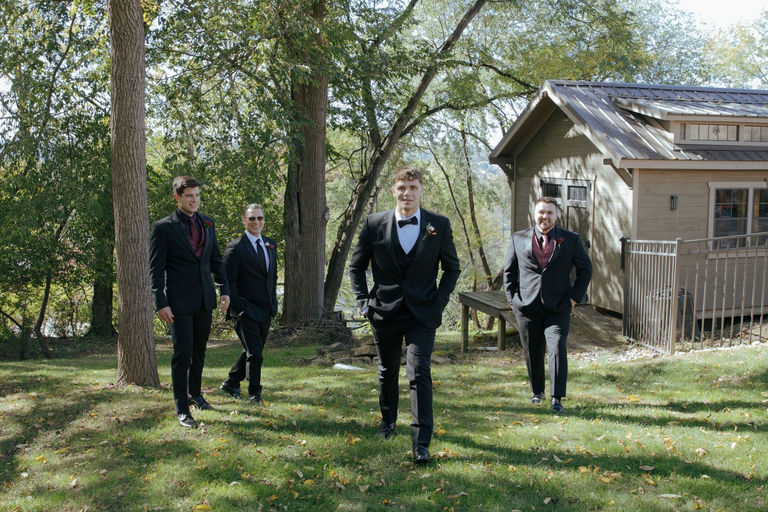 Four groomsmen in black tuxedos walking outdoors near a rustic wooden building surrounded by trees.