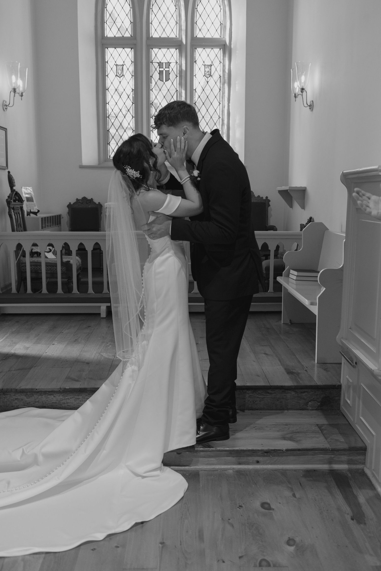 Bride and groom sharing first kiss in black and white photo inside a chapel with stained glass windows.
