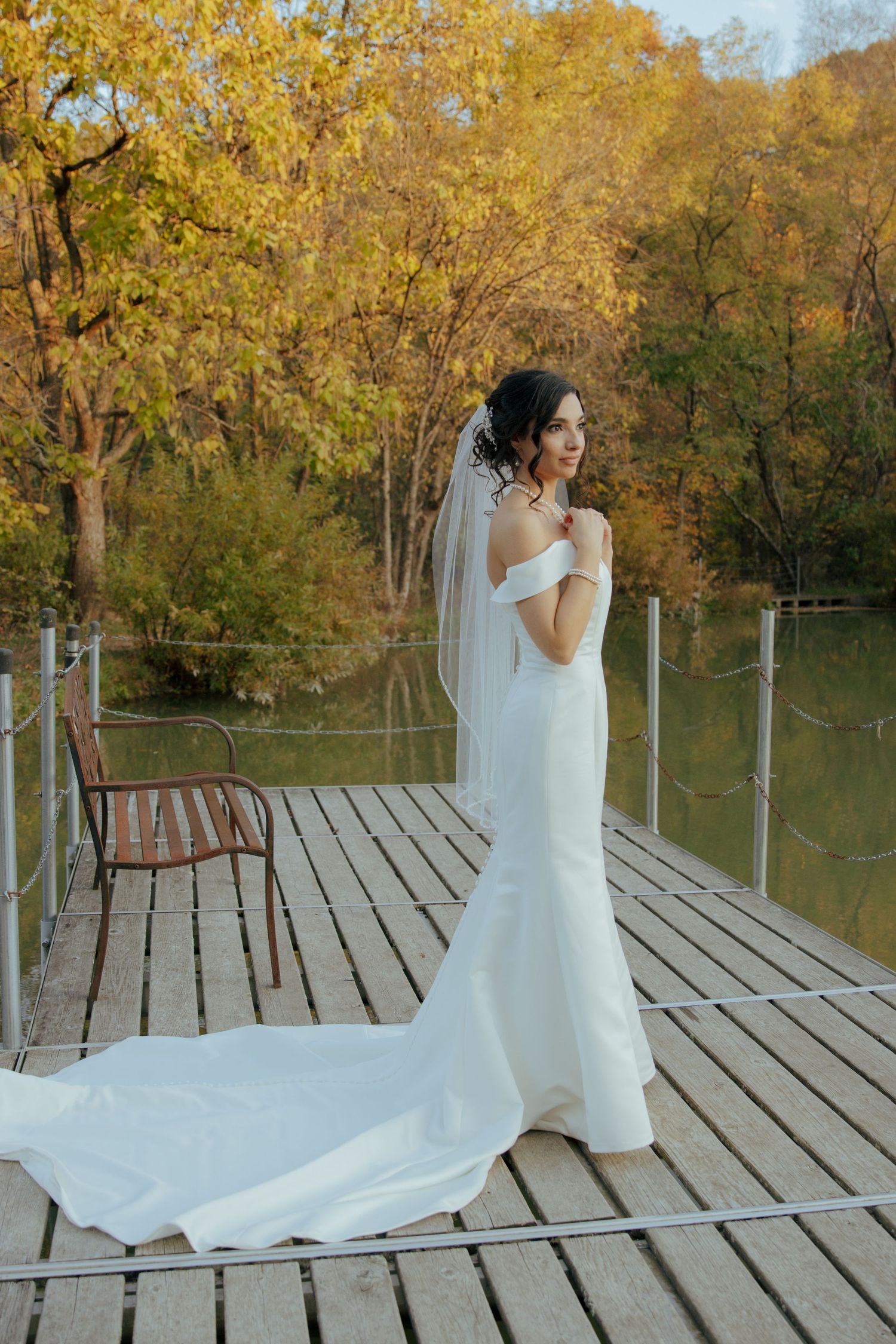 Bride in white off-shoulder gown with veil stands on wooden dock surrounded by autumn foliage and lake.