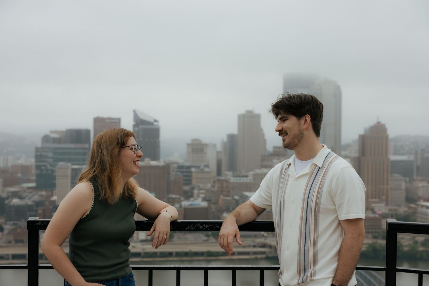 Couple smiling at each other on a rooftop overlooking a foggy city skyline.