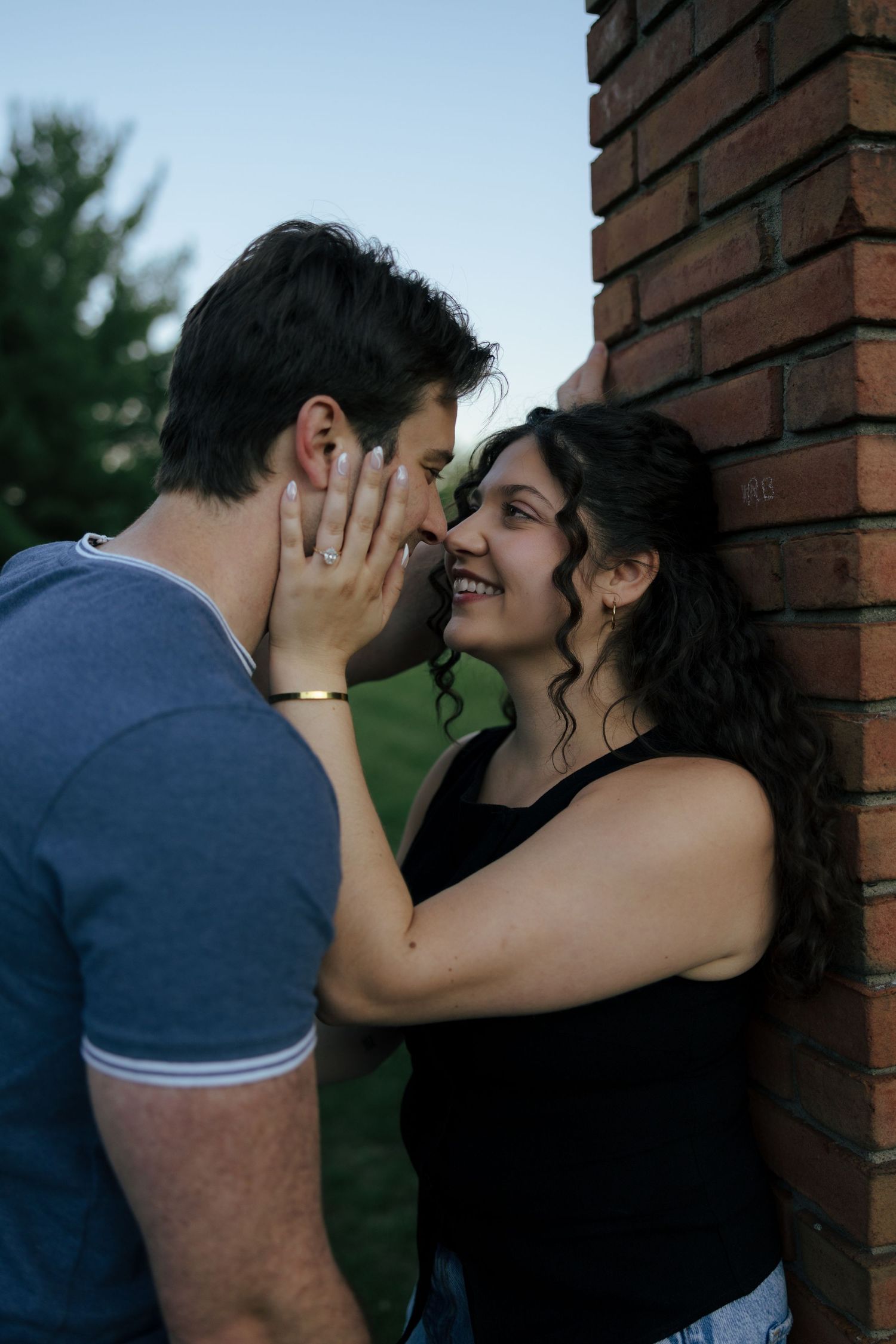 Couple sharing a romantic moment near a brick wall, woman smiling as she touches mans face outdoors.