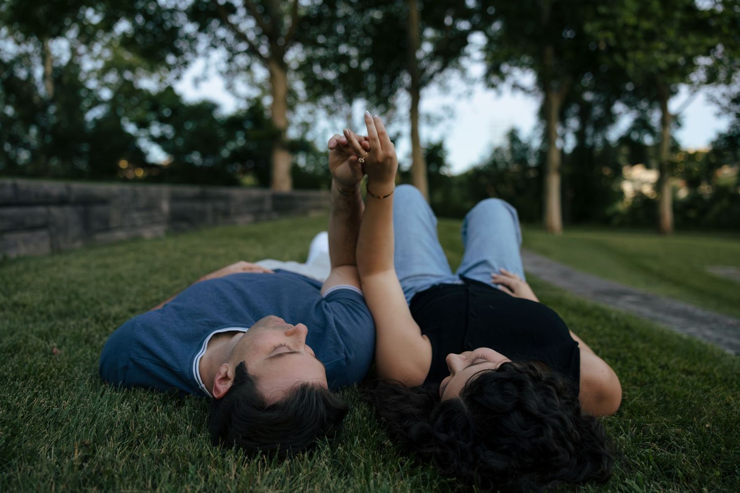 Couple lying on green grass in a park, holding hands and gazing at each other romantically at dusk.