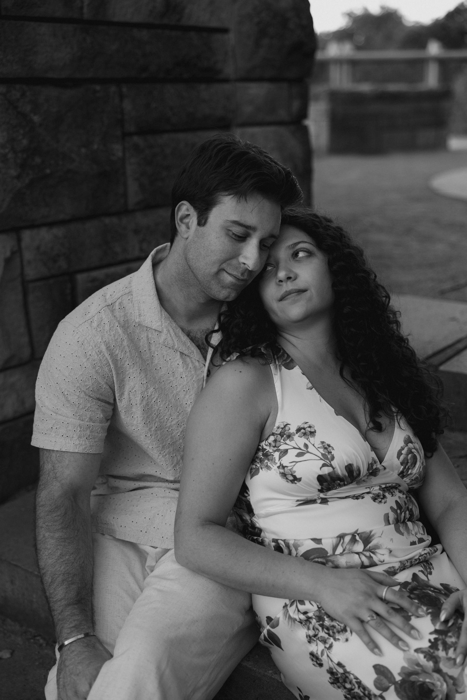 Black and white portrait of couple sitting together against a stone wall, woman leaning on man.