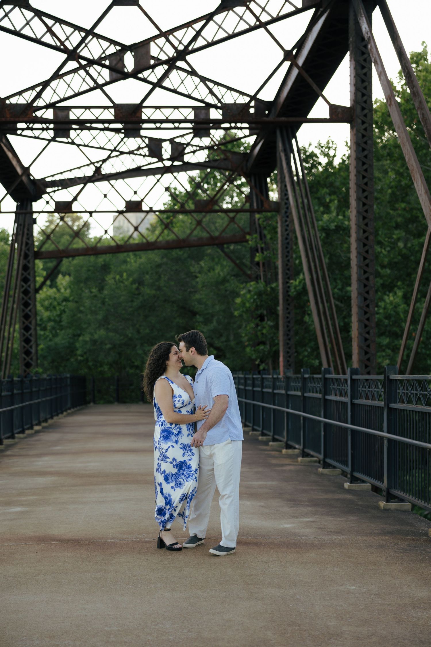 A couple shares a romantic kiss on a rustic iron bridge surrounded by lush green trees.