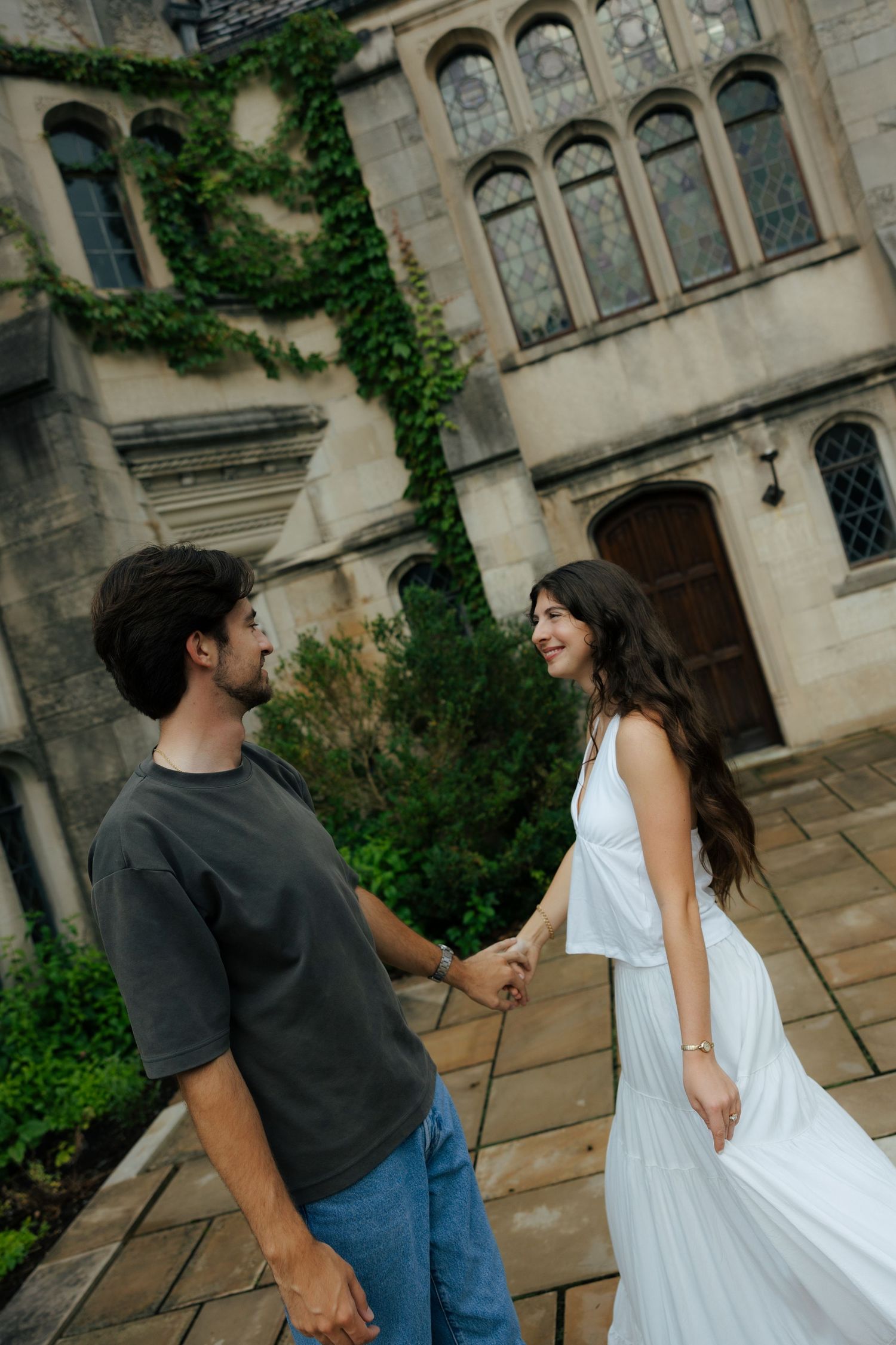 Couple holding hands outside ivy-covered stone building, woman in white dress smiling at partner.
