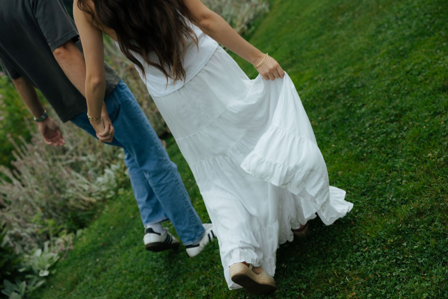 Couple walking on green grass, woman in flowing white wedding dress holding skirt, man in jeans and gray shirt holding her hand.