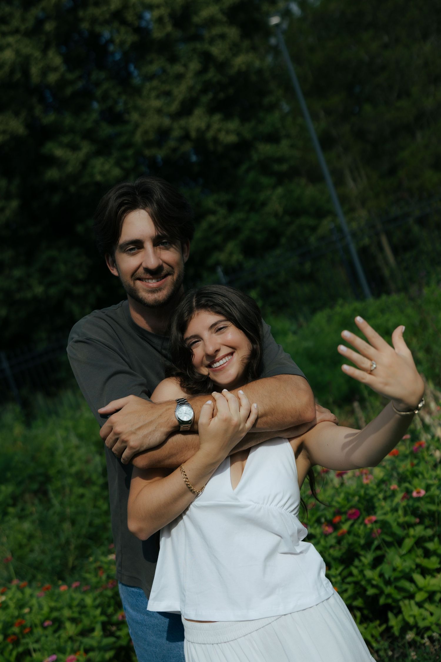 Smiling couple embracing outdoors, woman showing engagement ring in a lush garden.