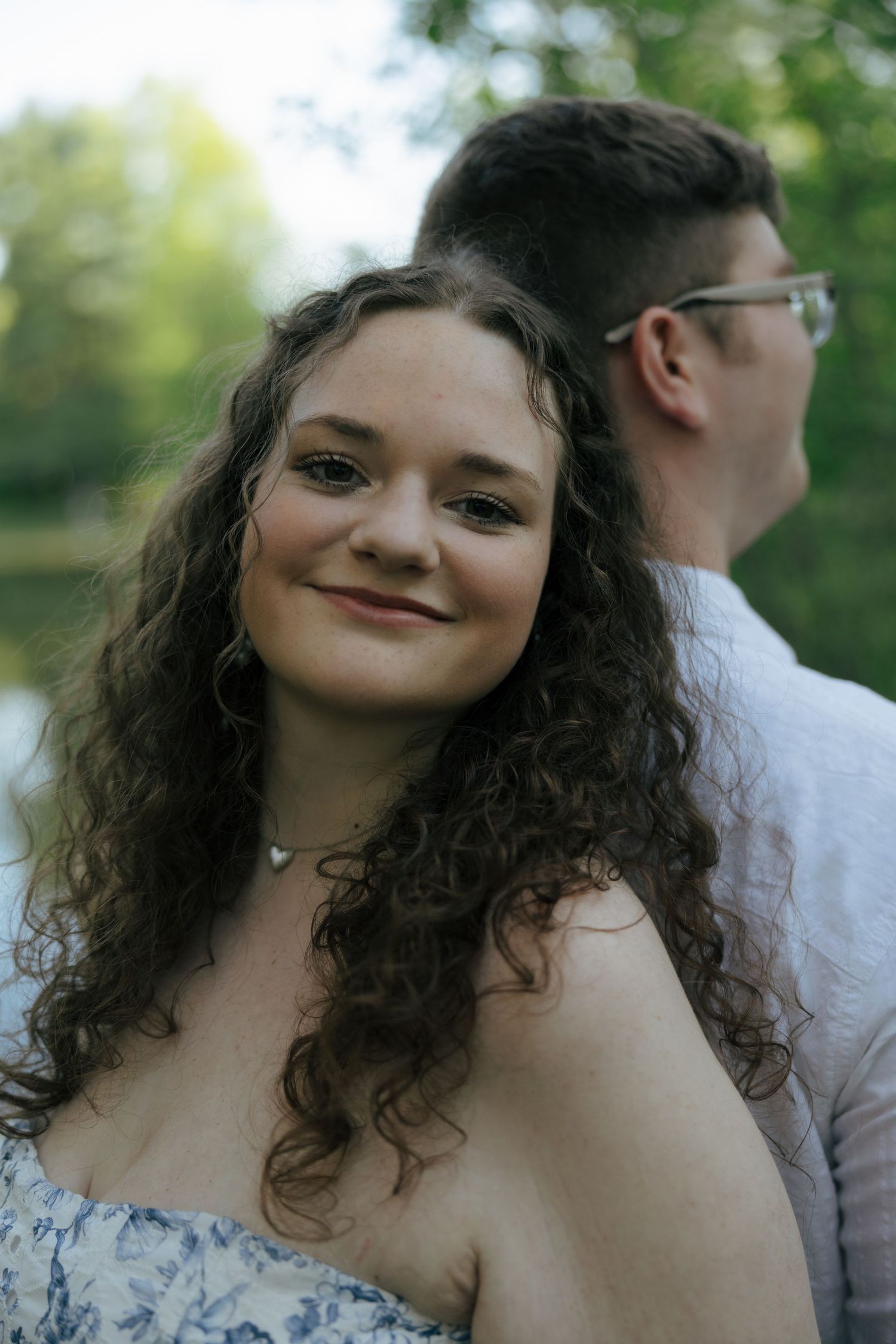 Young woman with curly brown hair in floral dress smiles at camera with her partner standing behind her outdoors.