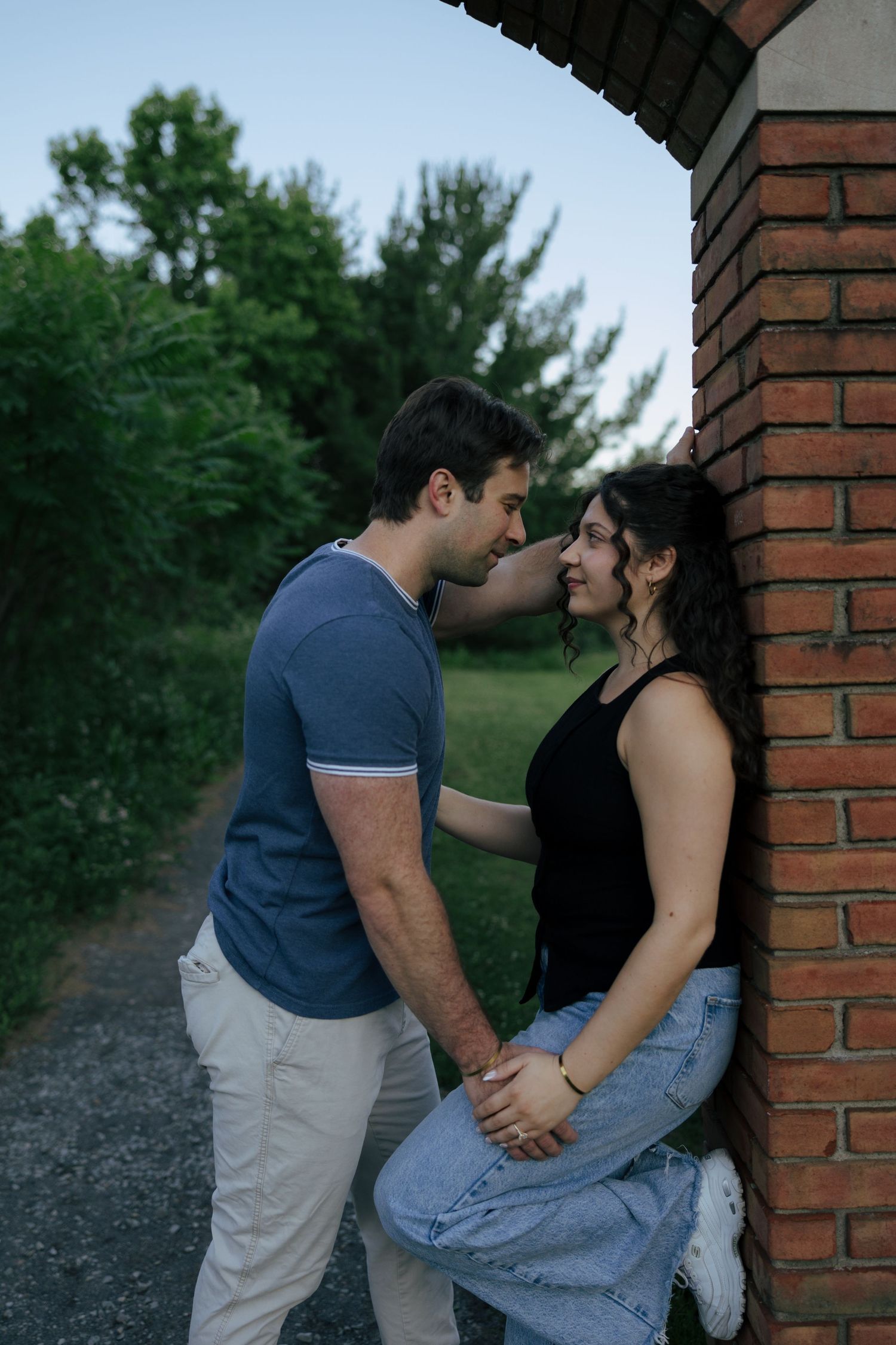 A young couple shares a romantic moment leaning against a brick wall outdoors, surrounded by lush green trees.
