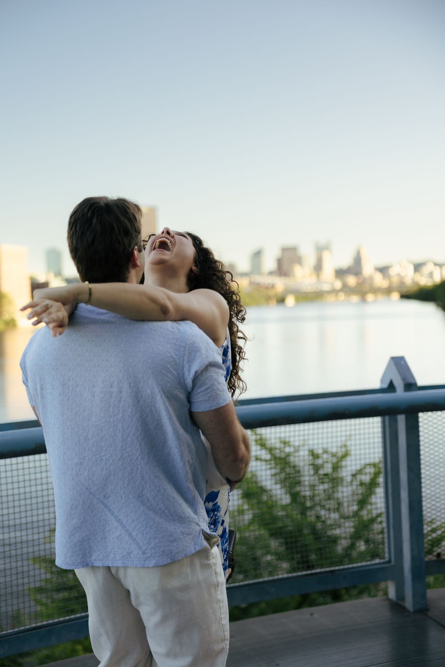 A joyful couple embraces on a riverside bridge, with a city skyline and clear blue sky in the background.