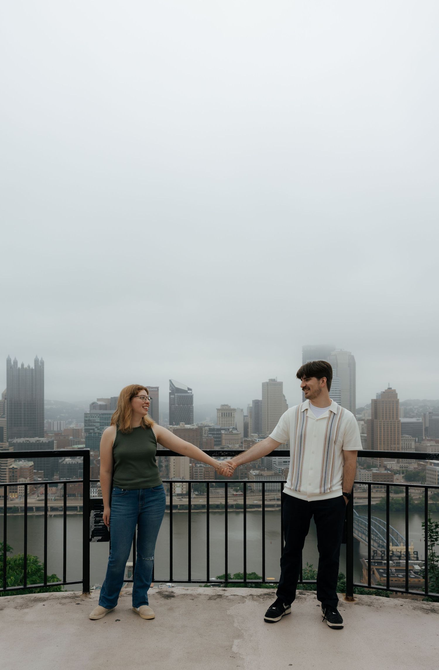 Couple holds hands on overlook with Pittsburgh skyline in misty background.