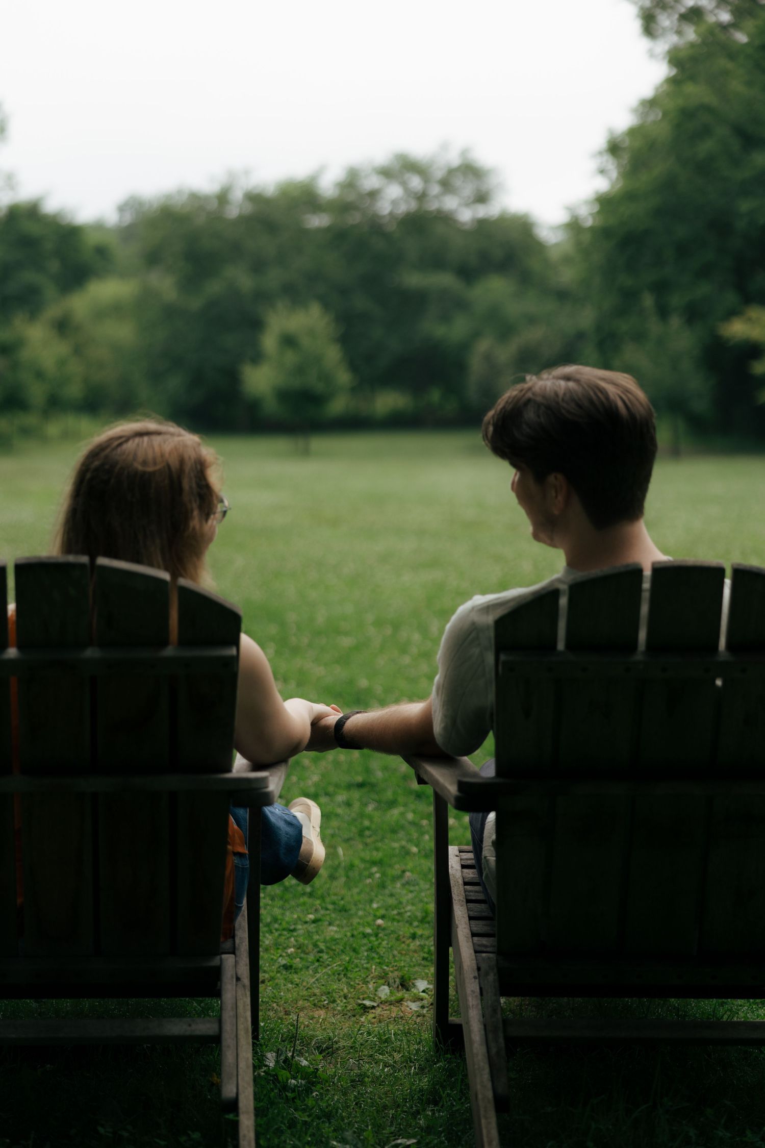 Couple holding hands while seated in wooden Adirondack chairs overlooking a green park lawn.