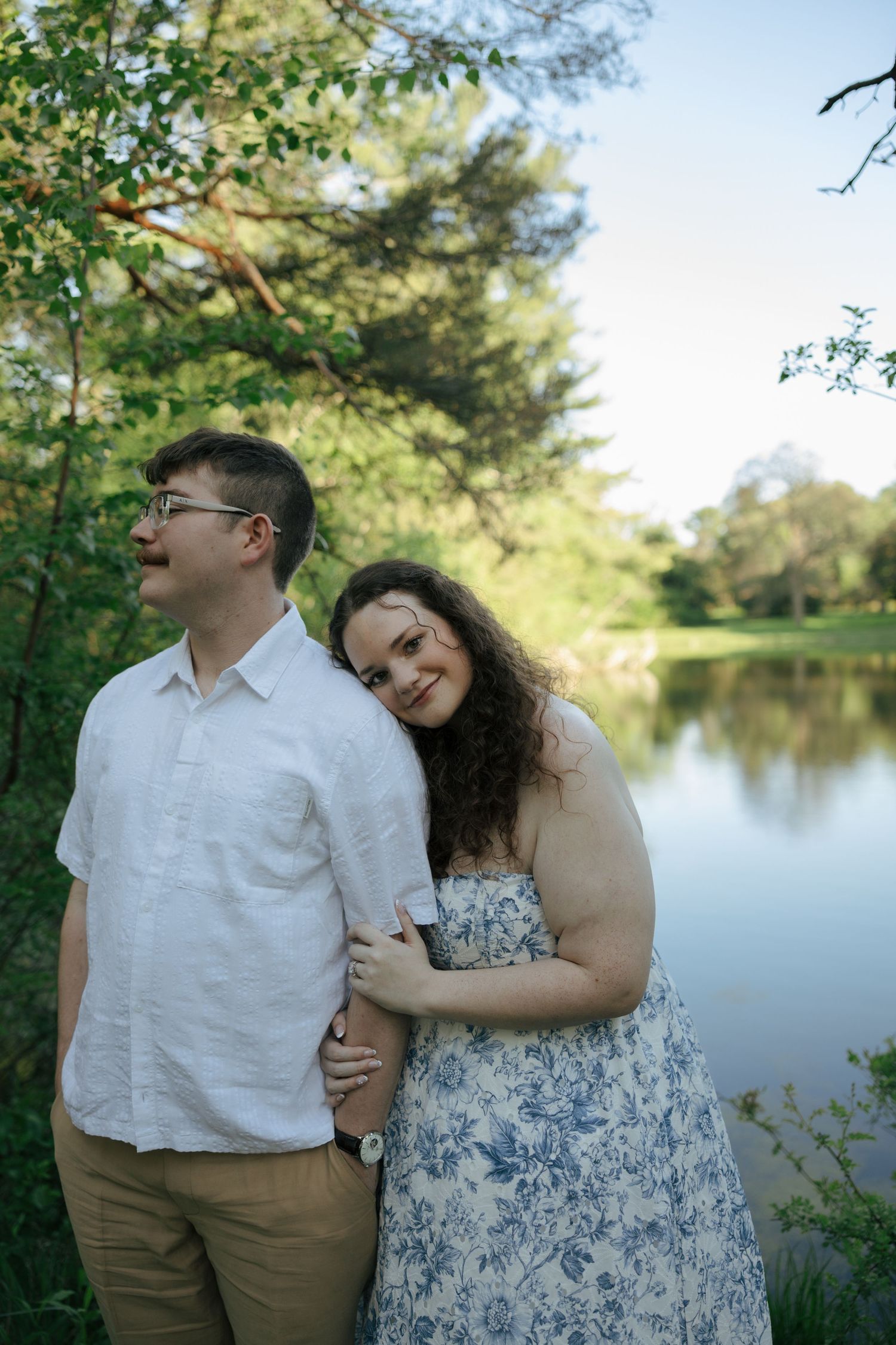 Couple embracing near a serene lake surrounded by lush green trees on a sunny day.