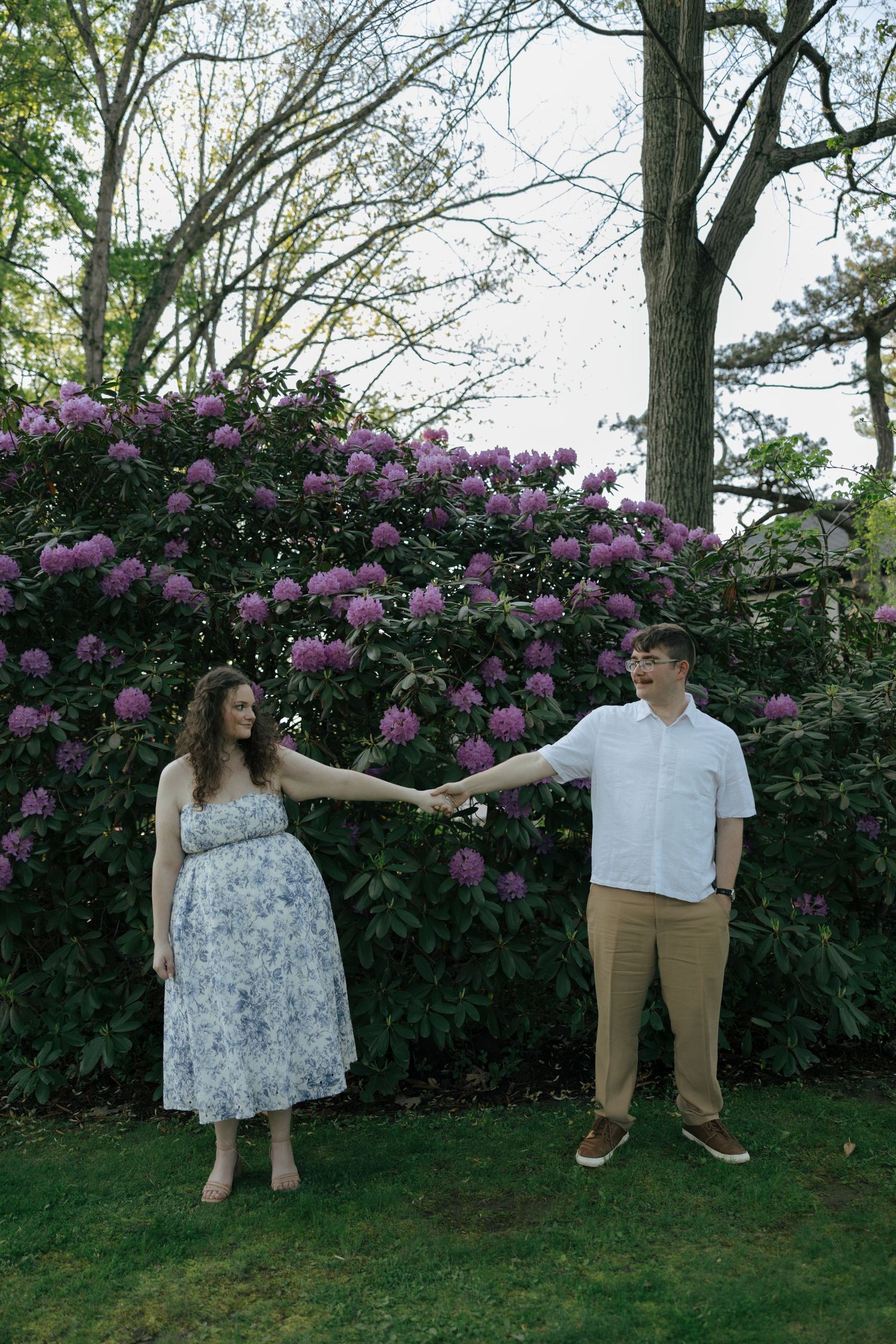 Engaged couple holding hands at arm's length in front of vibrant purple rhododendron bushes in a lush garden.