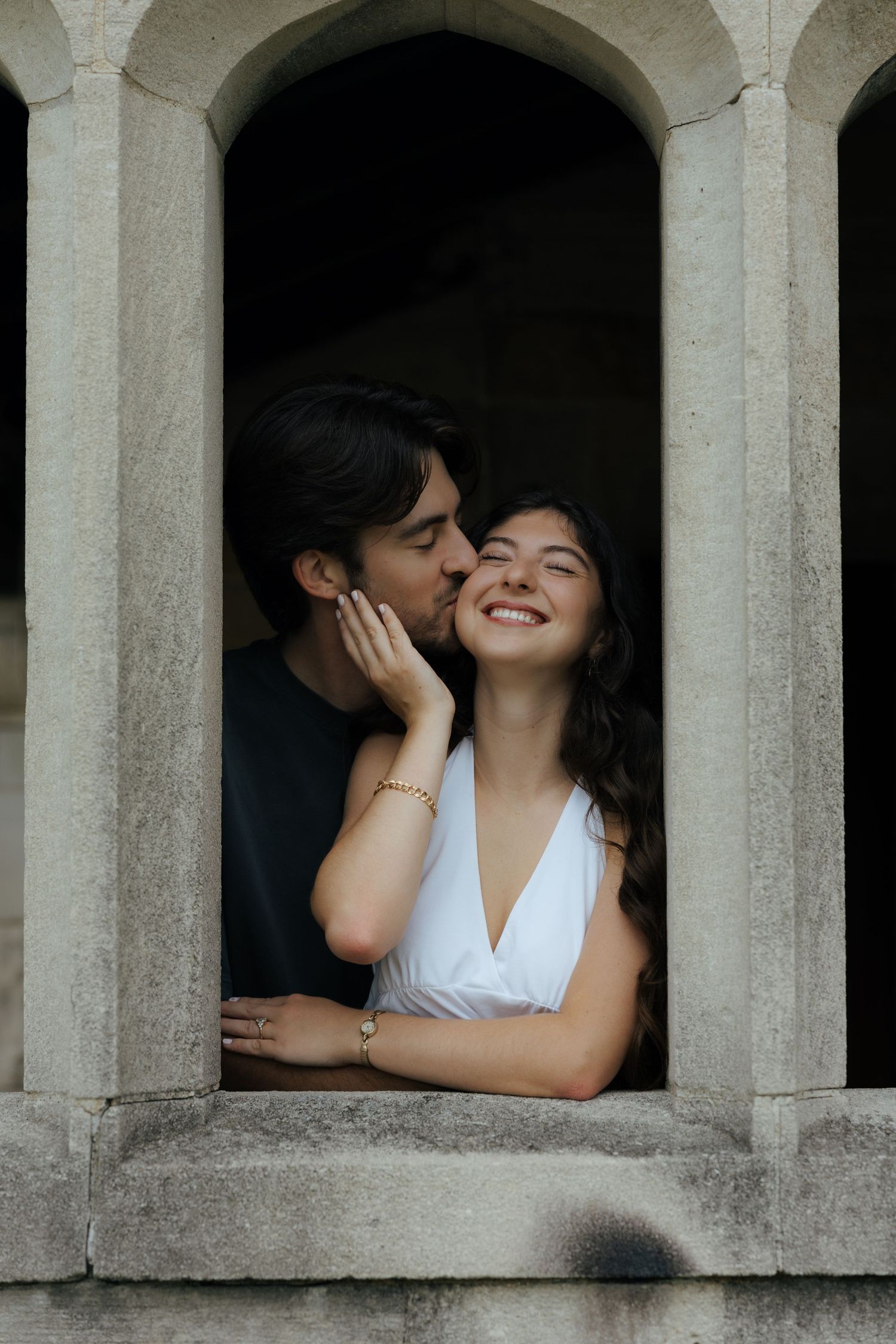 A romantic couple shares a tender kiss in an arched stone window, the woman smiling in a white dress.