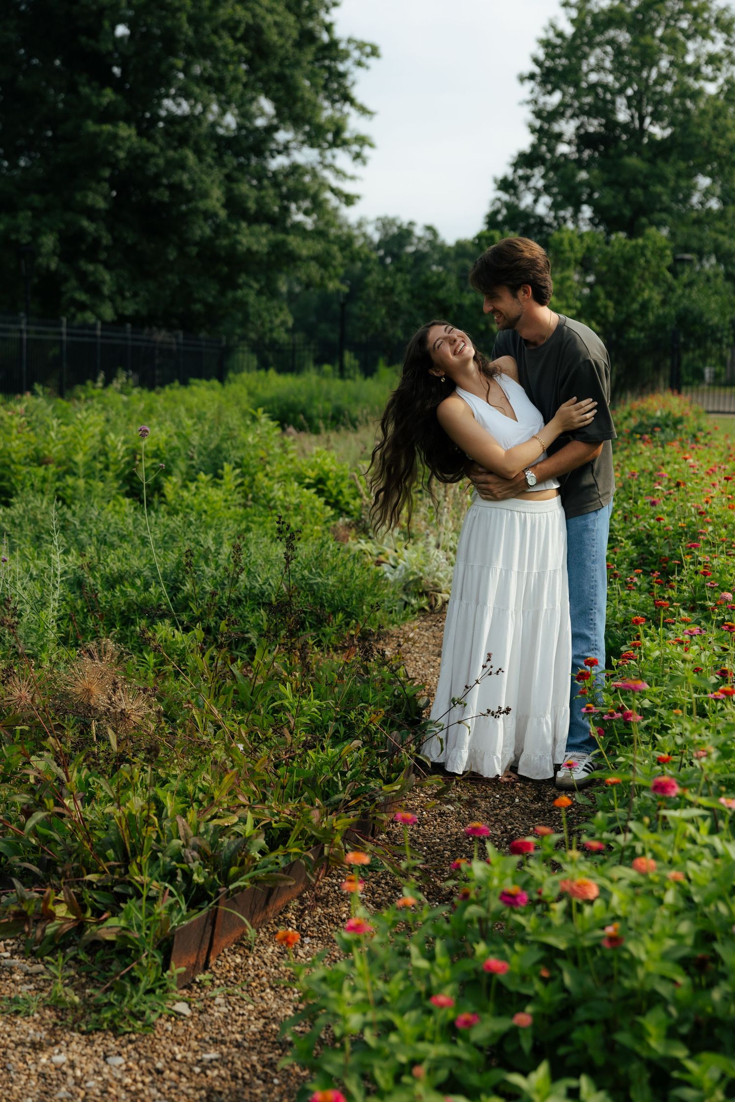Couple embracing and laughing together along a garden path lined with colorful zinnia flowers.