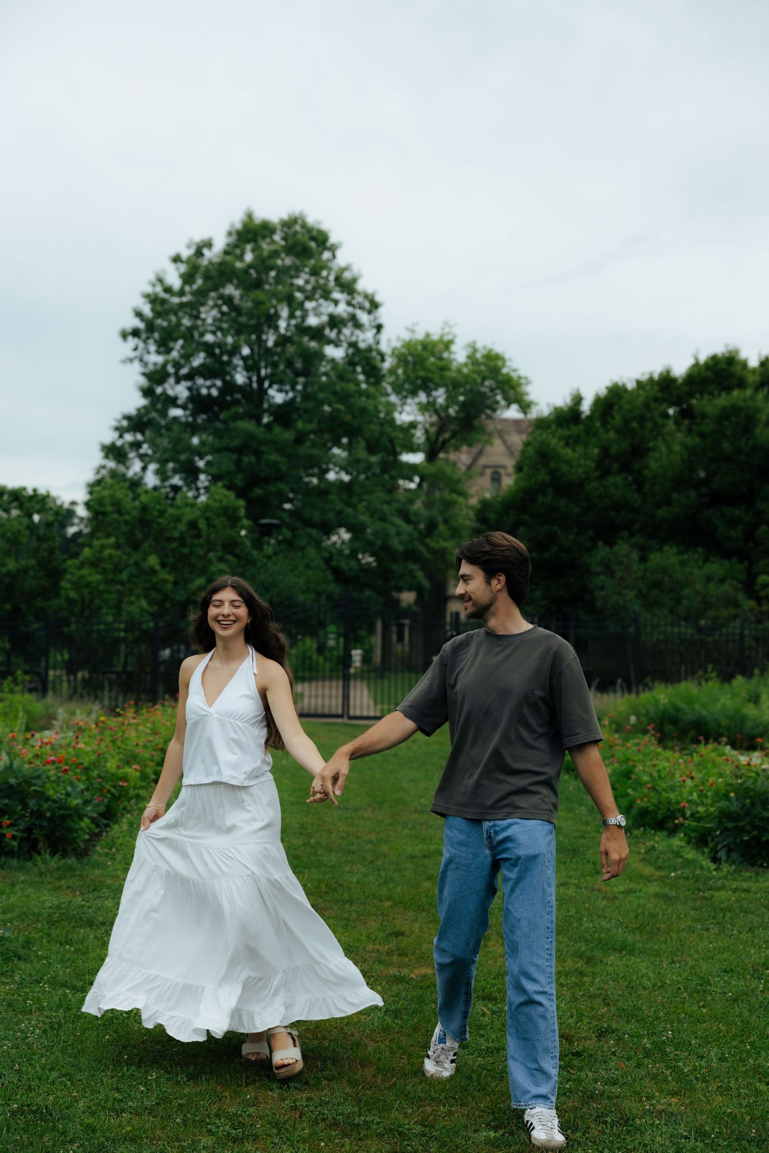 A smiling couple holds hands while walking through a lush green park, woman in white dress, man in gray tee and jeans.
