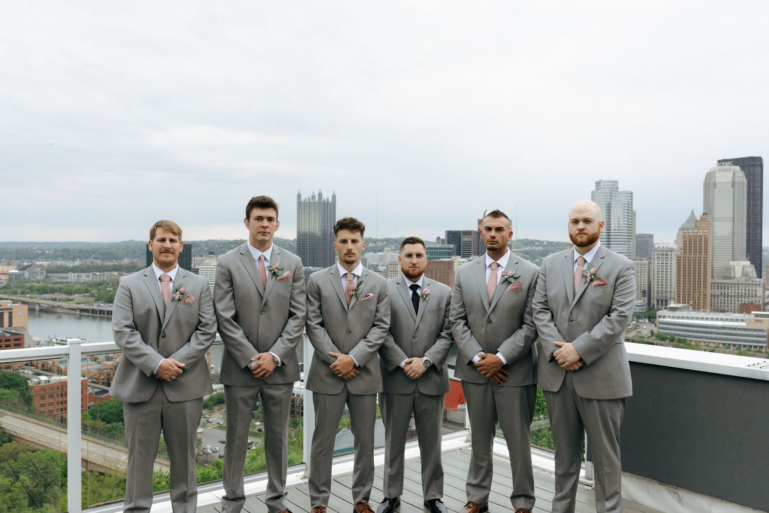 Six groomsmen in gray suits pose on a rooftop with the Pittsburgh city skyline in the background.