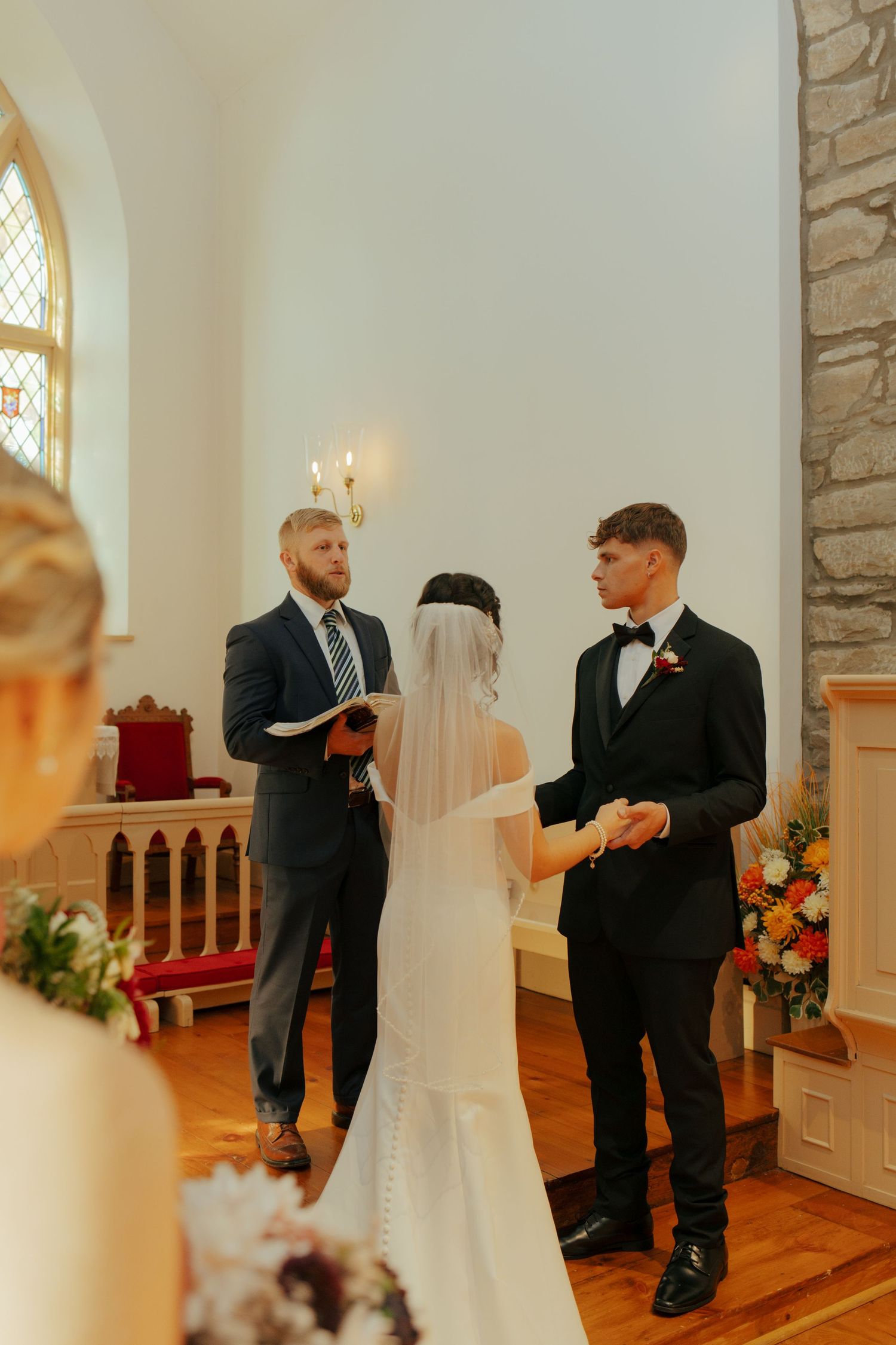 Bride and groom exchange vows at church altar with officiant holding open book during wedding ceremony.