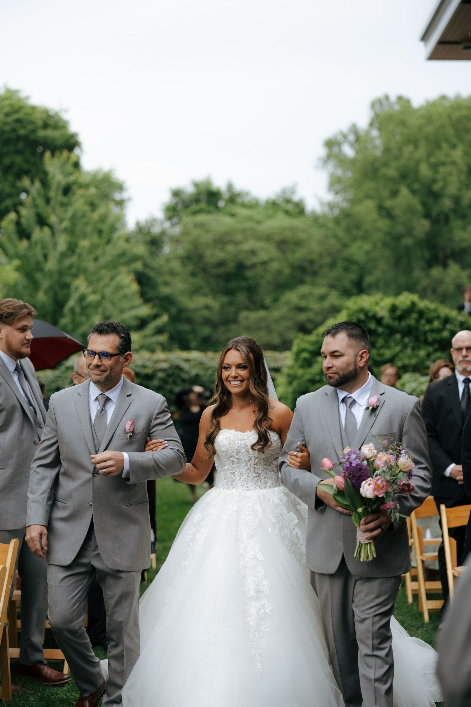 Bride in white ballgown walks down outdoor aisle with two groomsmen in gray suits holding purple bouquet.