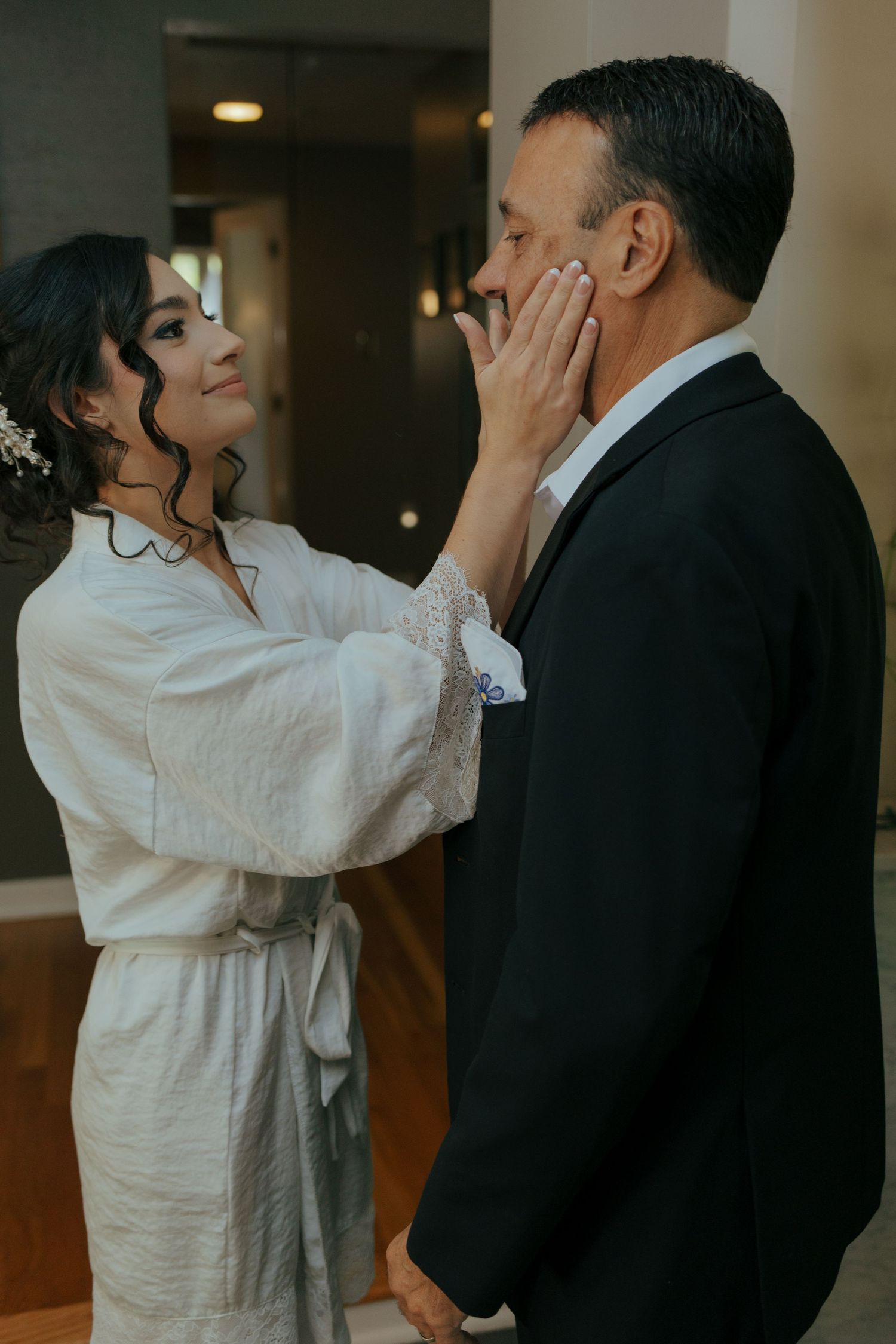 Bride in white robe tenderly touches grooms face during an emotional first look moment indoors.