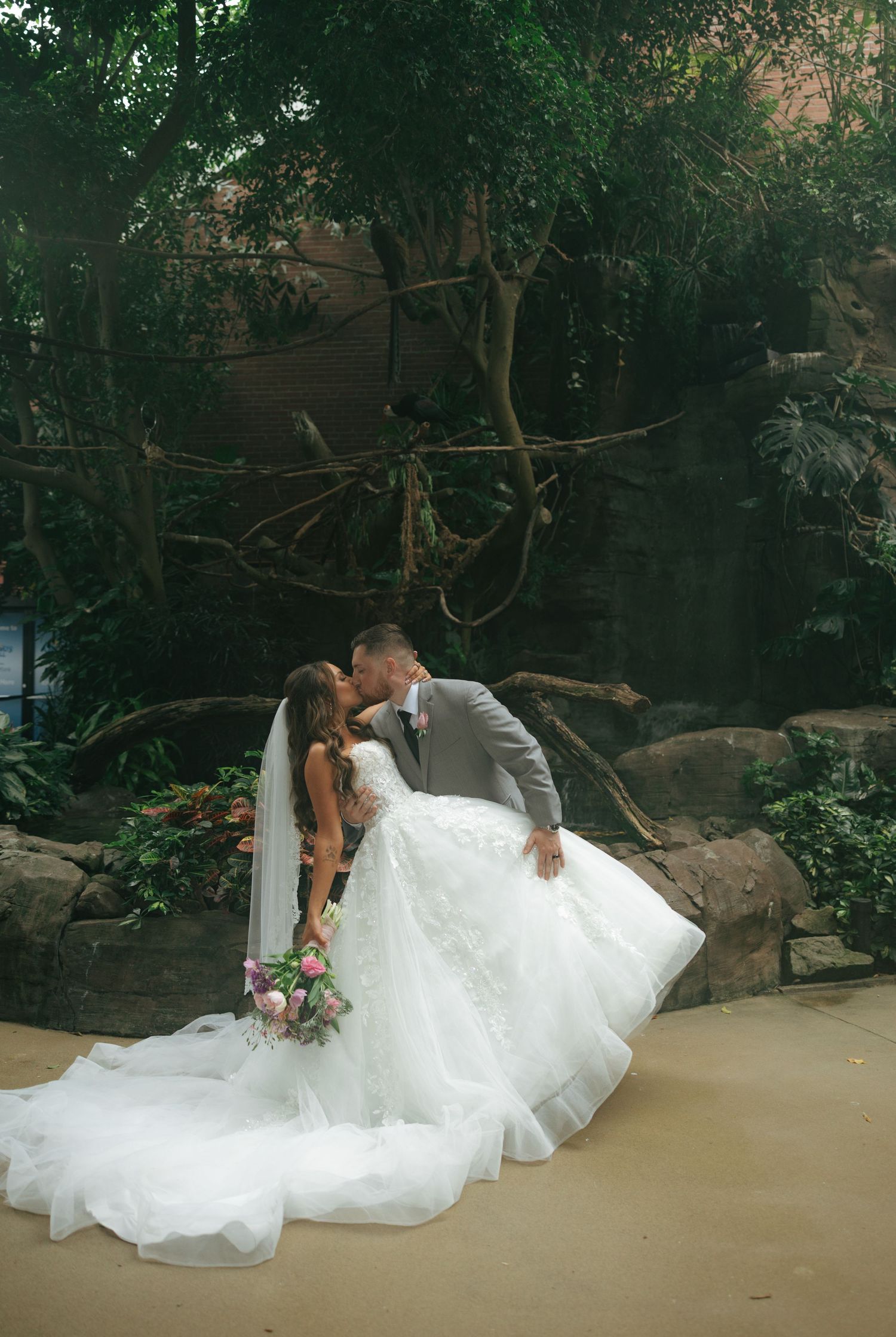 Bride and groom share a romantic kiss in a lush garden setting, bride wearing a flowing white ballgown with long veil.