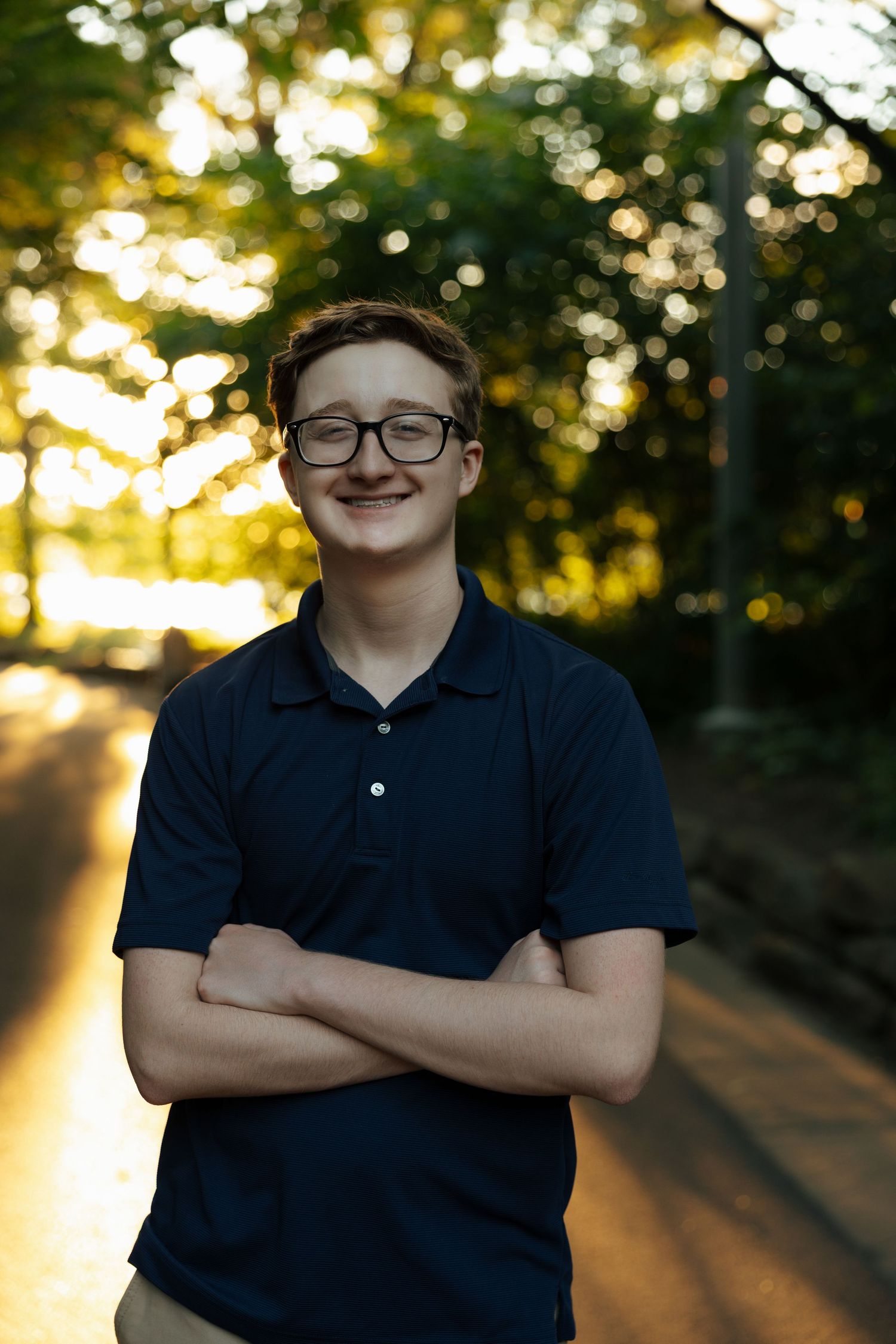 Smiling teenage boy with glasses and crossed arms wearing navy polo shirt outdoors at golden hour.