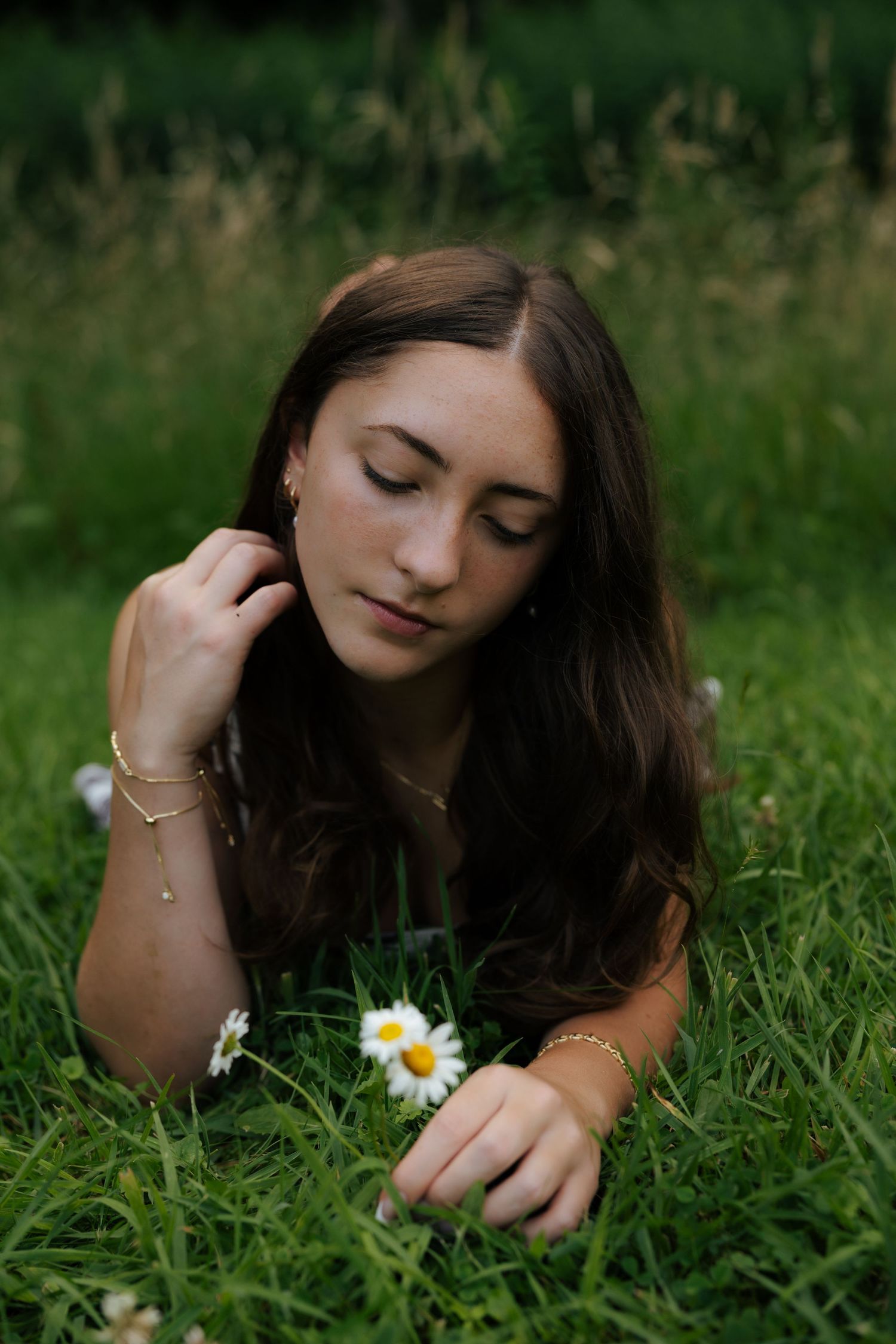 Young brunette woman lying in grass touching daisy flower, wearing gold jewelry outdoors.