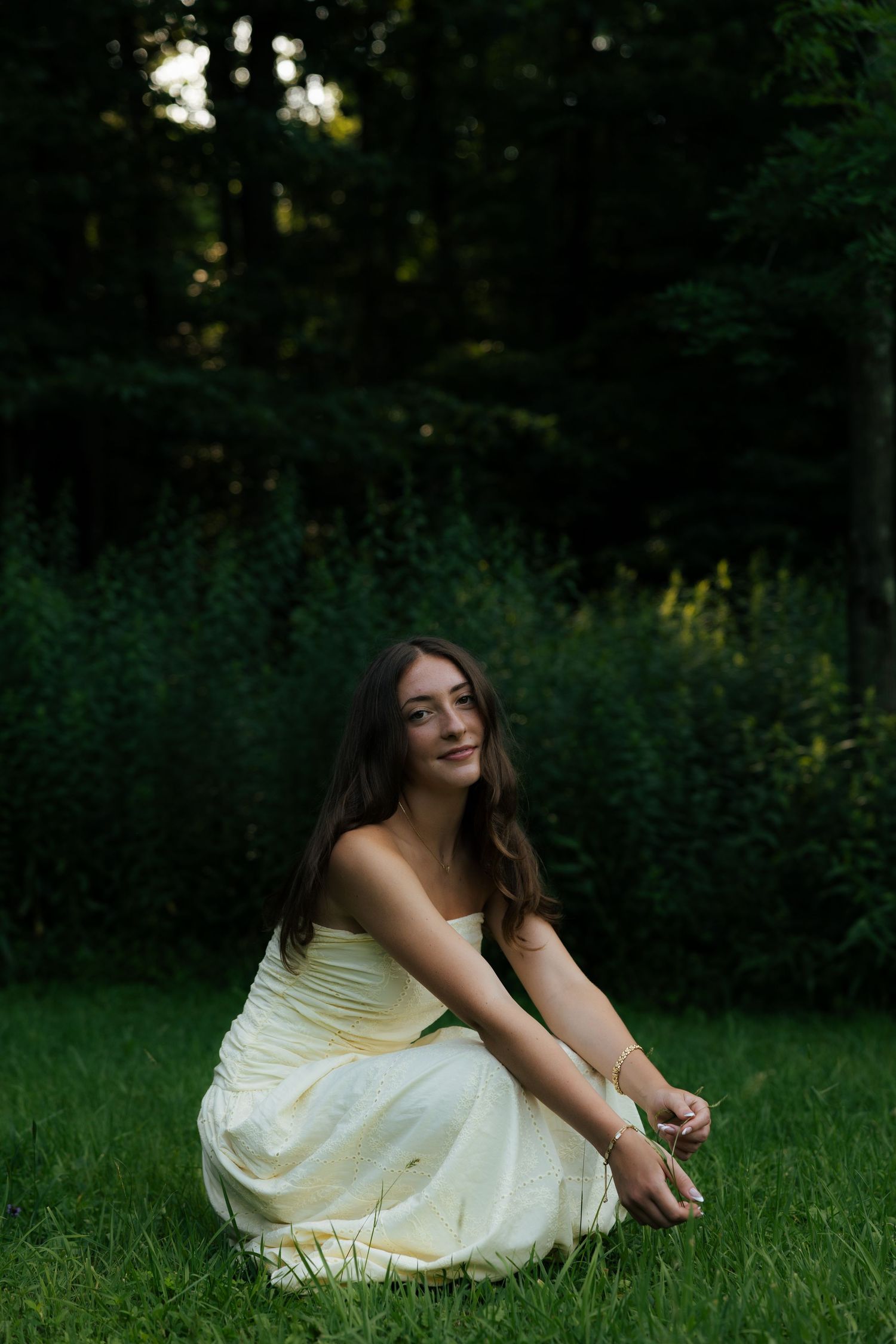 Young woman in yellow dress sitting on grass in lush green garden, smiling softly at camera.
