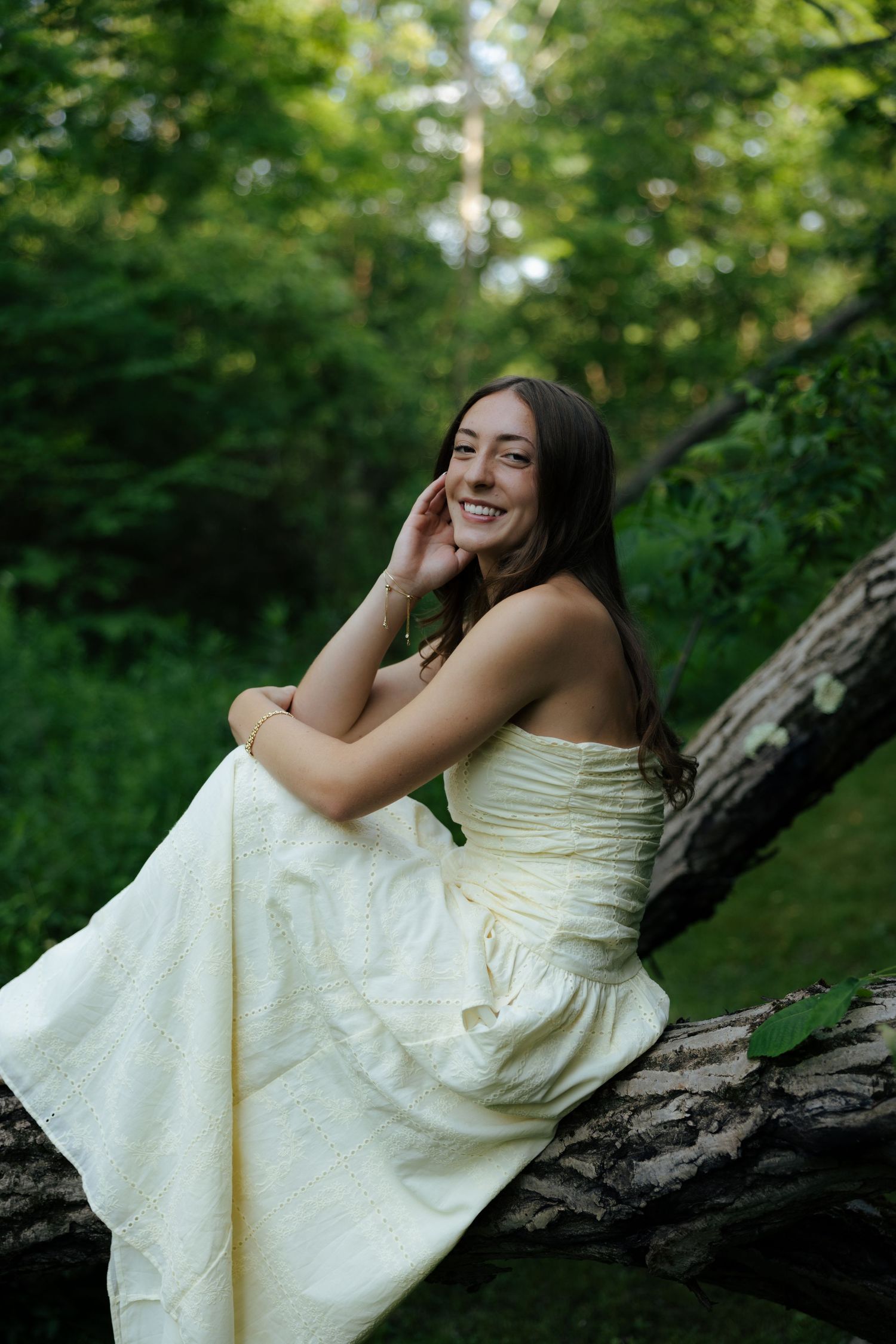 Young woman in white dress sitting on fallen log smiling in lush green forest setting.
