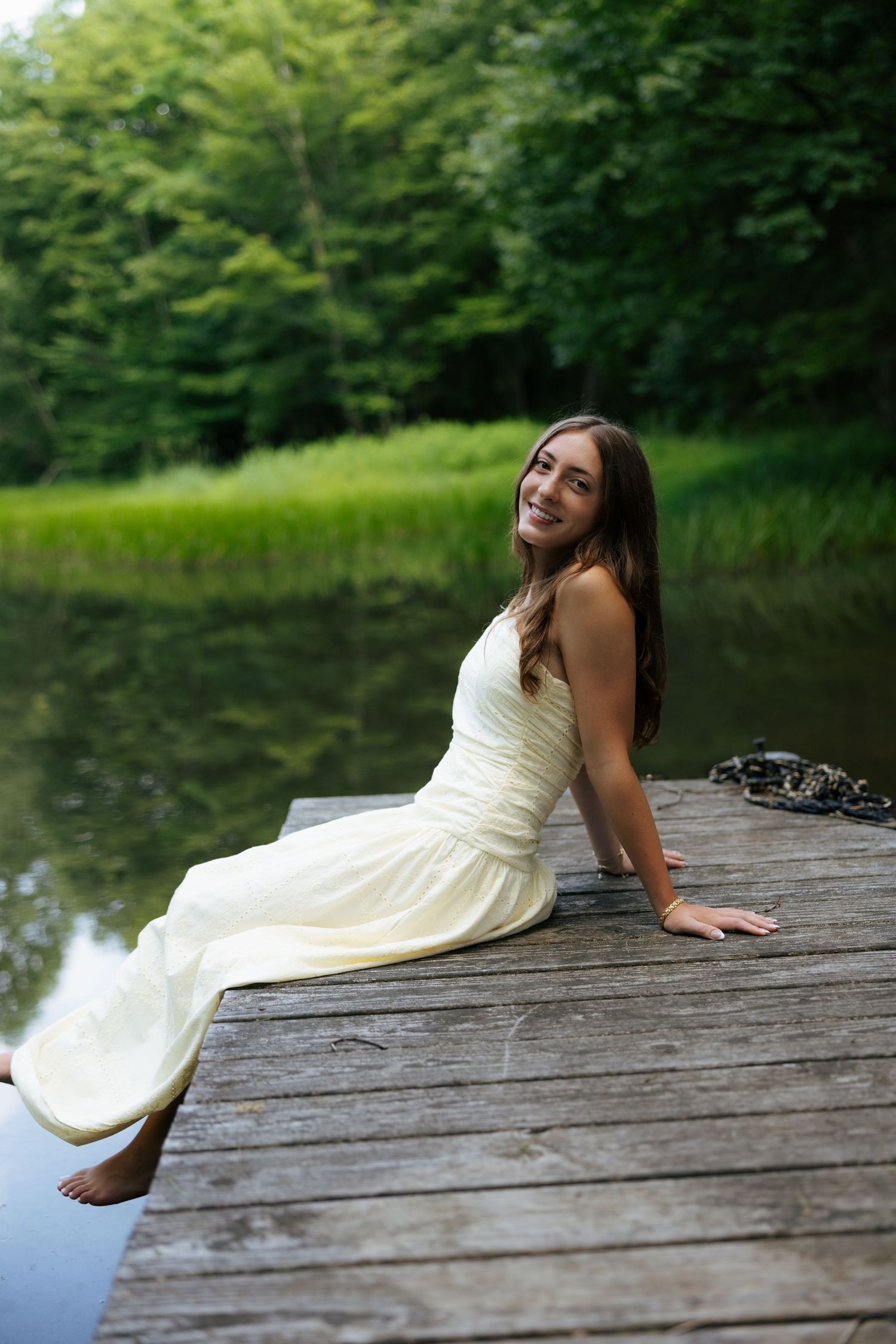 Smiling young woman in white dress sitting on wooden dock beside tranquil lake in woods.