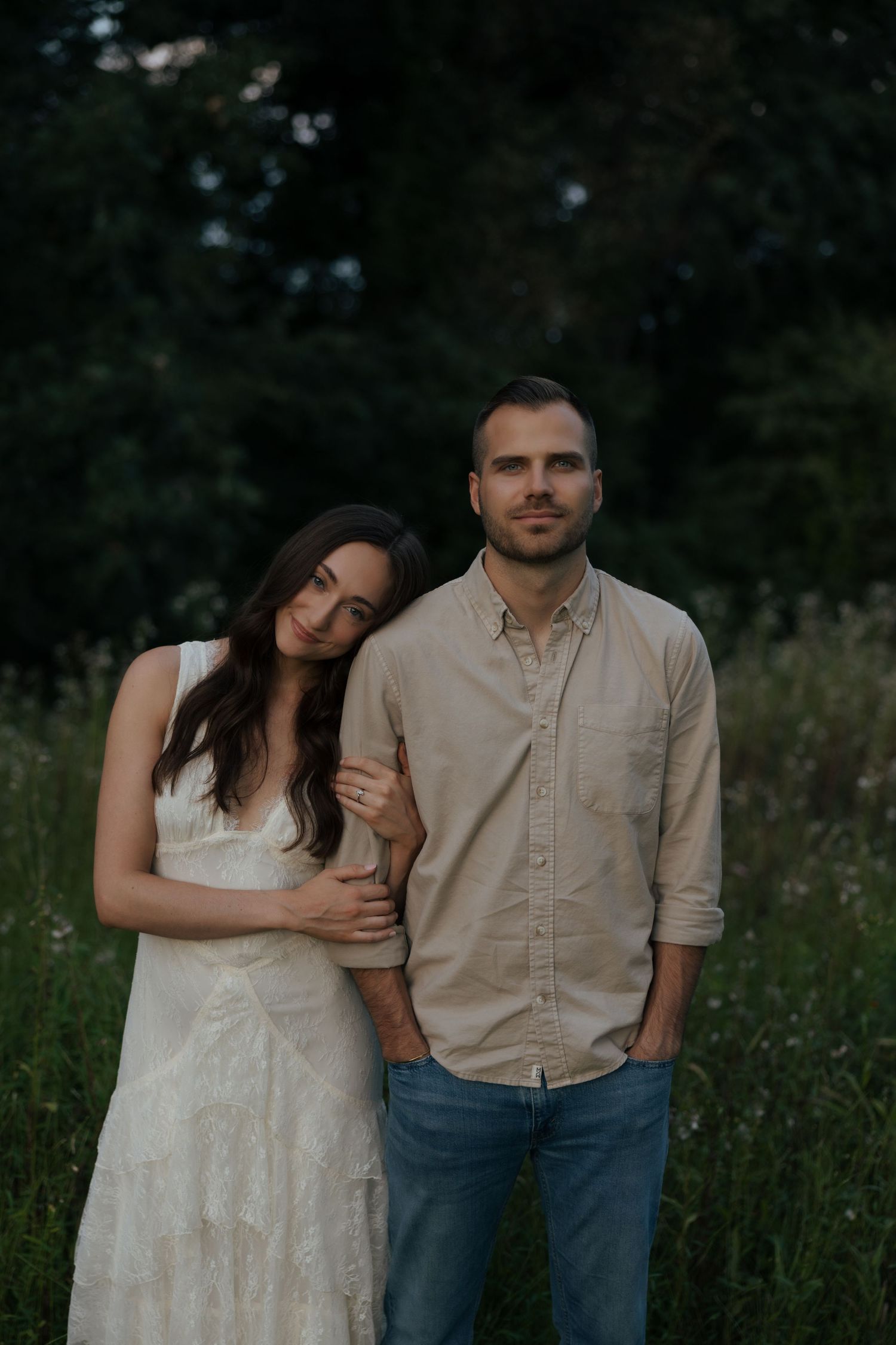 A couple poses together in a field, woman in white lace dress leaning on man in beige shirt.
