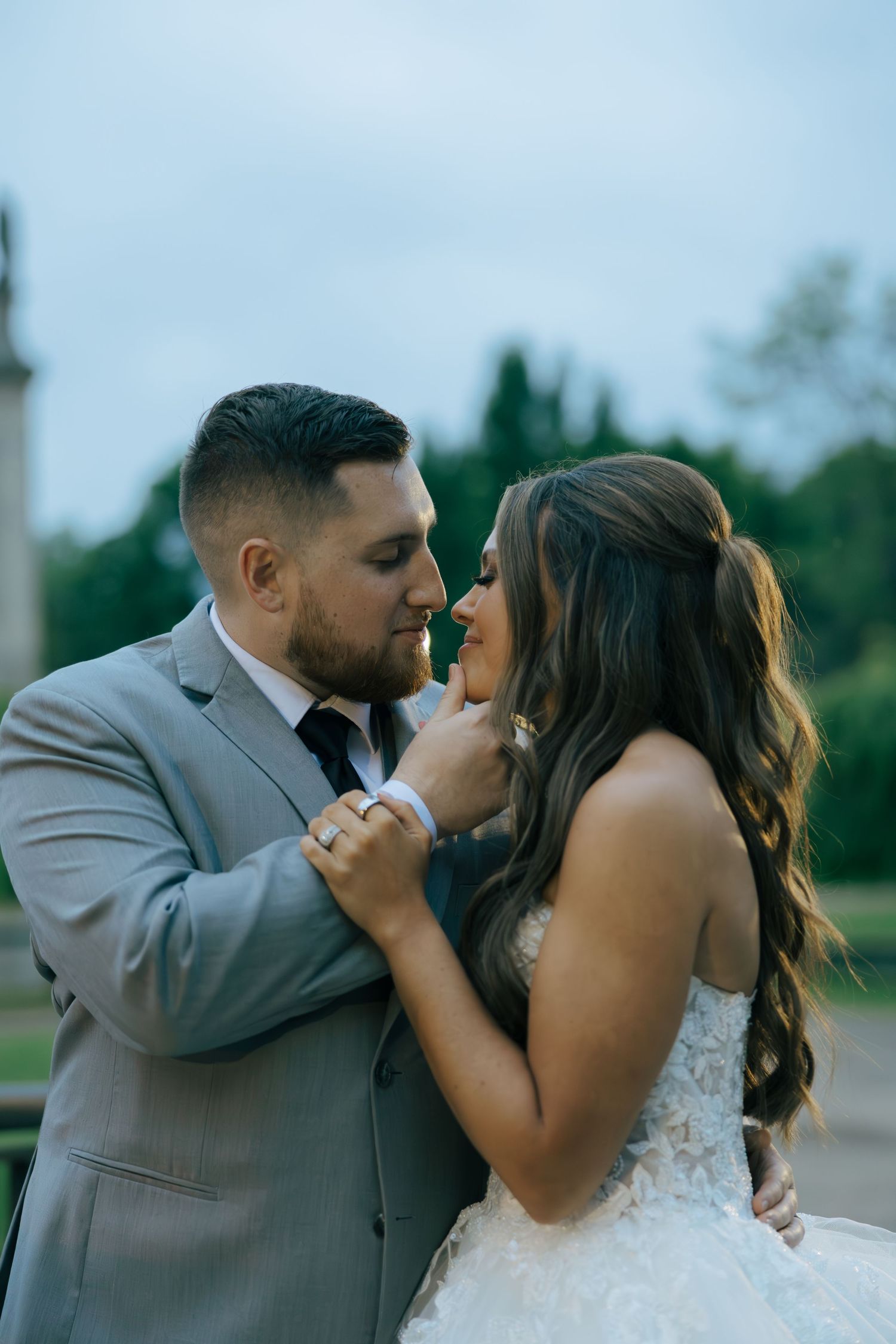 Groom in a gray suit and bride in a lace dress share a romantic moment outdoors at dusk.