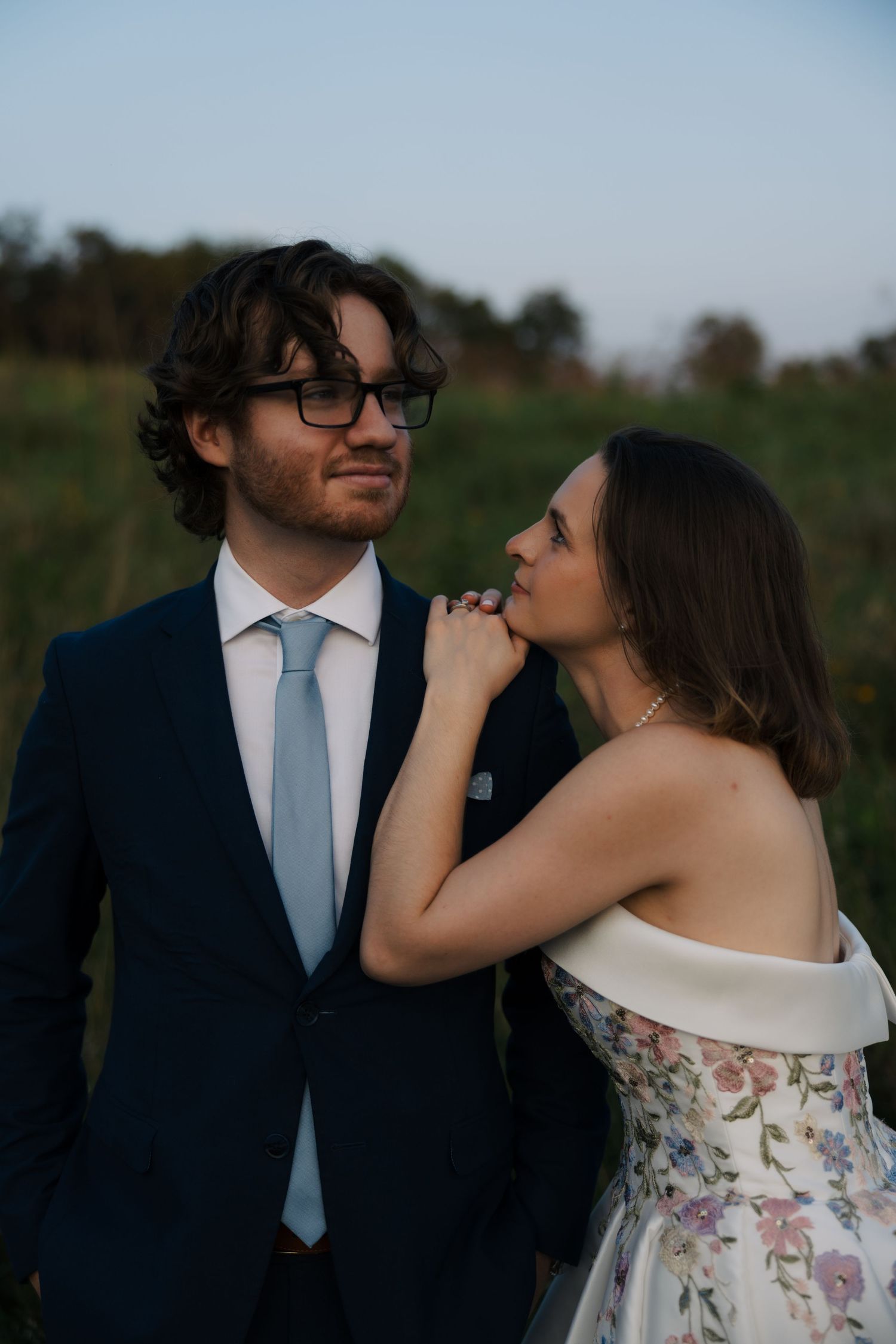 Bride in strapless floral gown gazes at groom in navy suit and glasses while holding his hand outdoors.