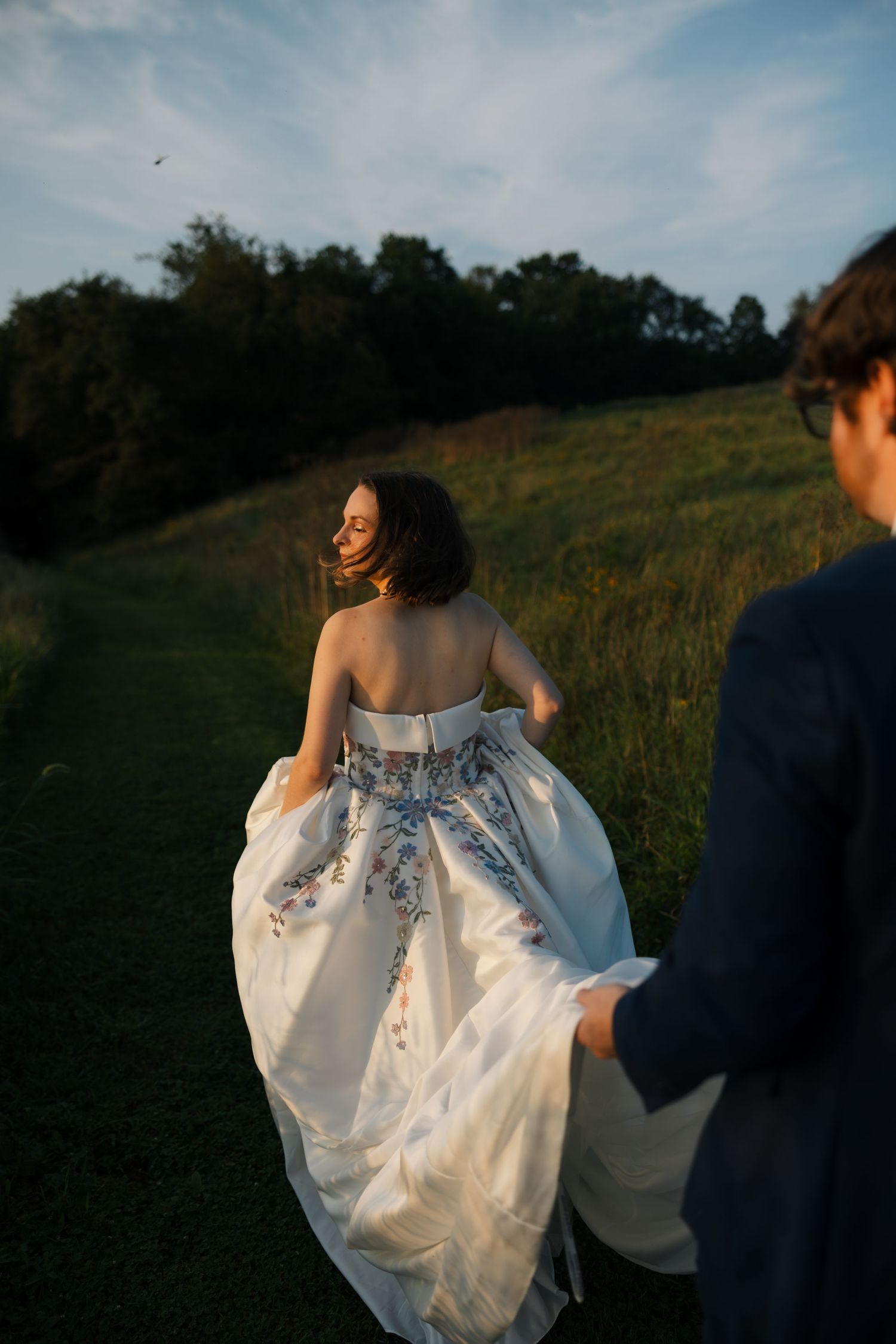 Bride in floral embroidered strapless gown walks with groom through green meadow at golden hour.