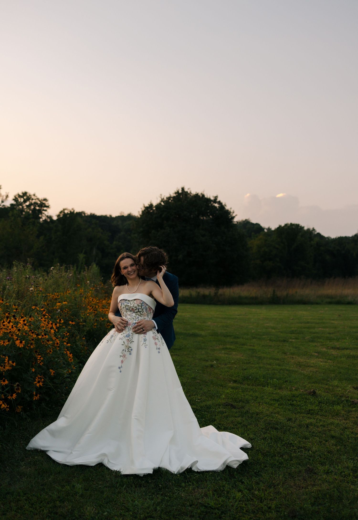 Bride in strapless floral ball gown with long train embraces groom near orange wildflowers at dusk.